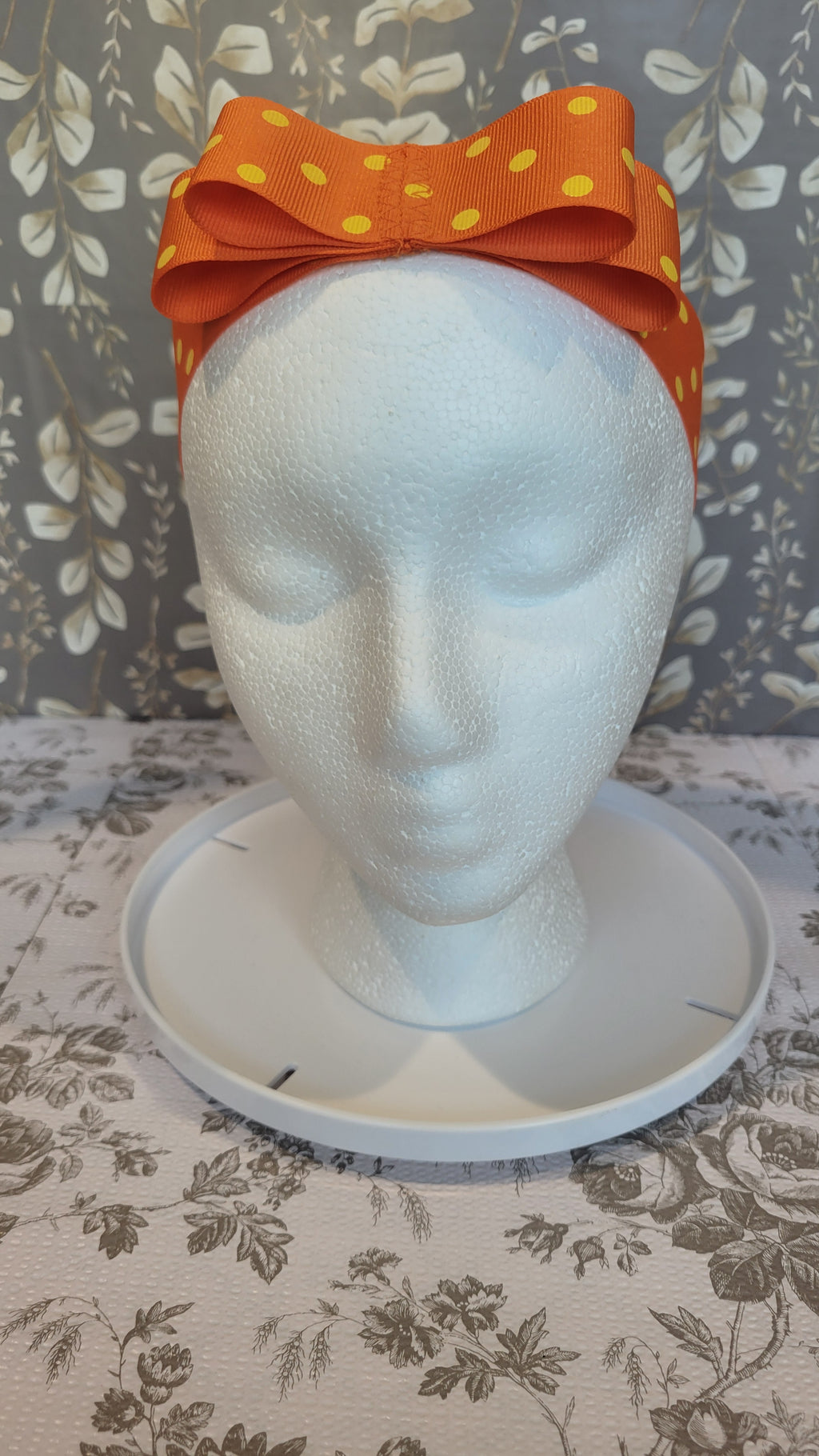 Mannequin head with orange polka dot bow on a hairband on a floral patterned wall