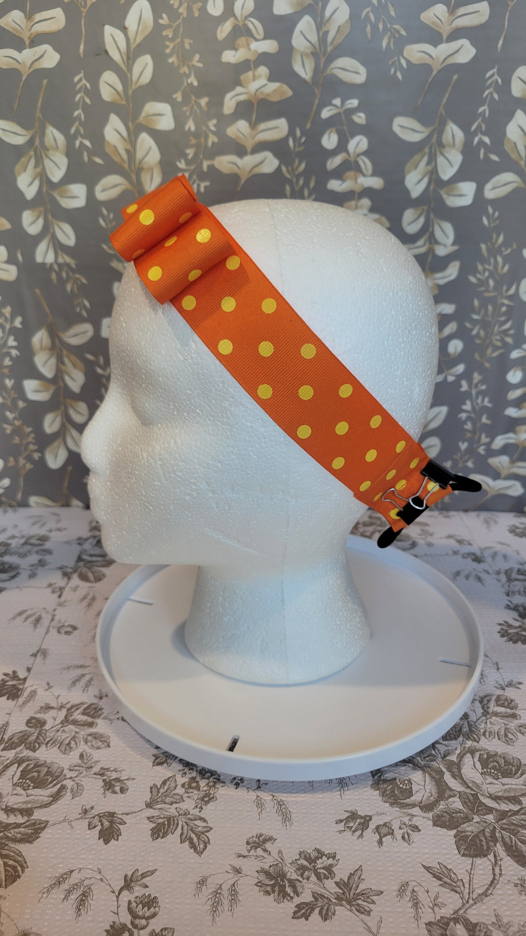 Mannequin head from the right side with orange polka dot bow on a hairband on a floral patterned wall 