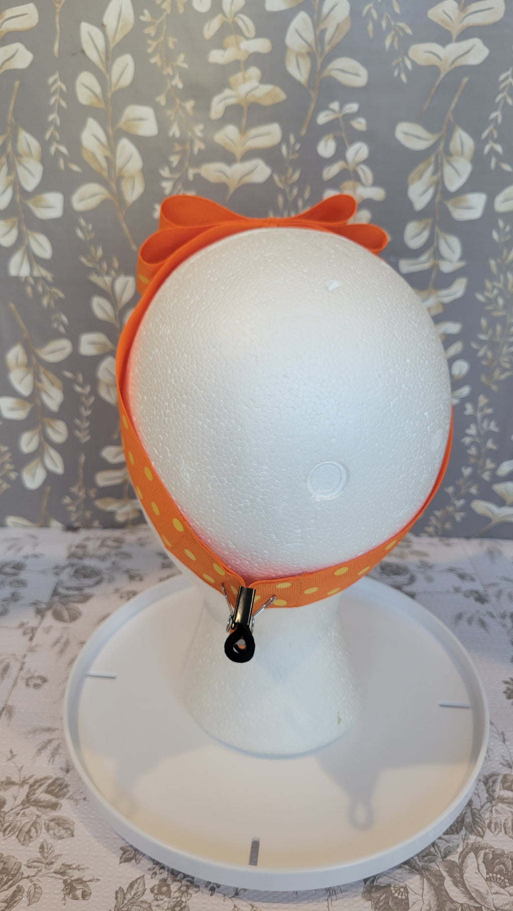 Mannequin head from the back with orange polka dot bow on a hairband on a floral patterned wall