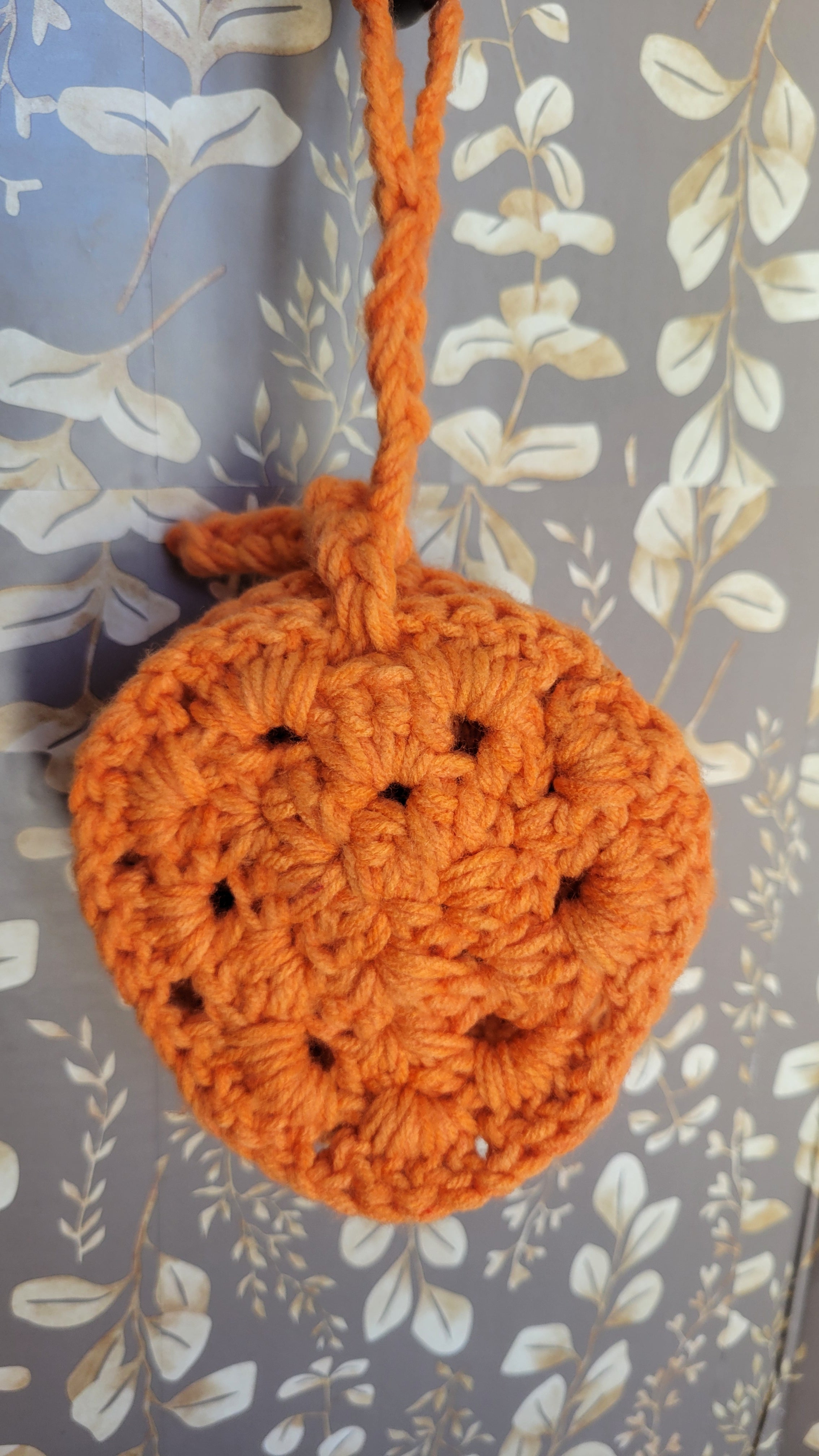 Four orange crocheted coasters hanging on a brown and tan leaf patterned surface