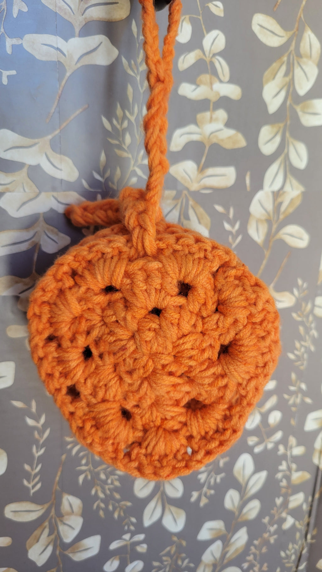Four orange crocheted coasters hanging on a brown and tan leaf patterned surface