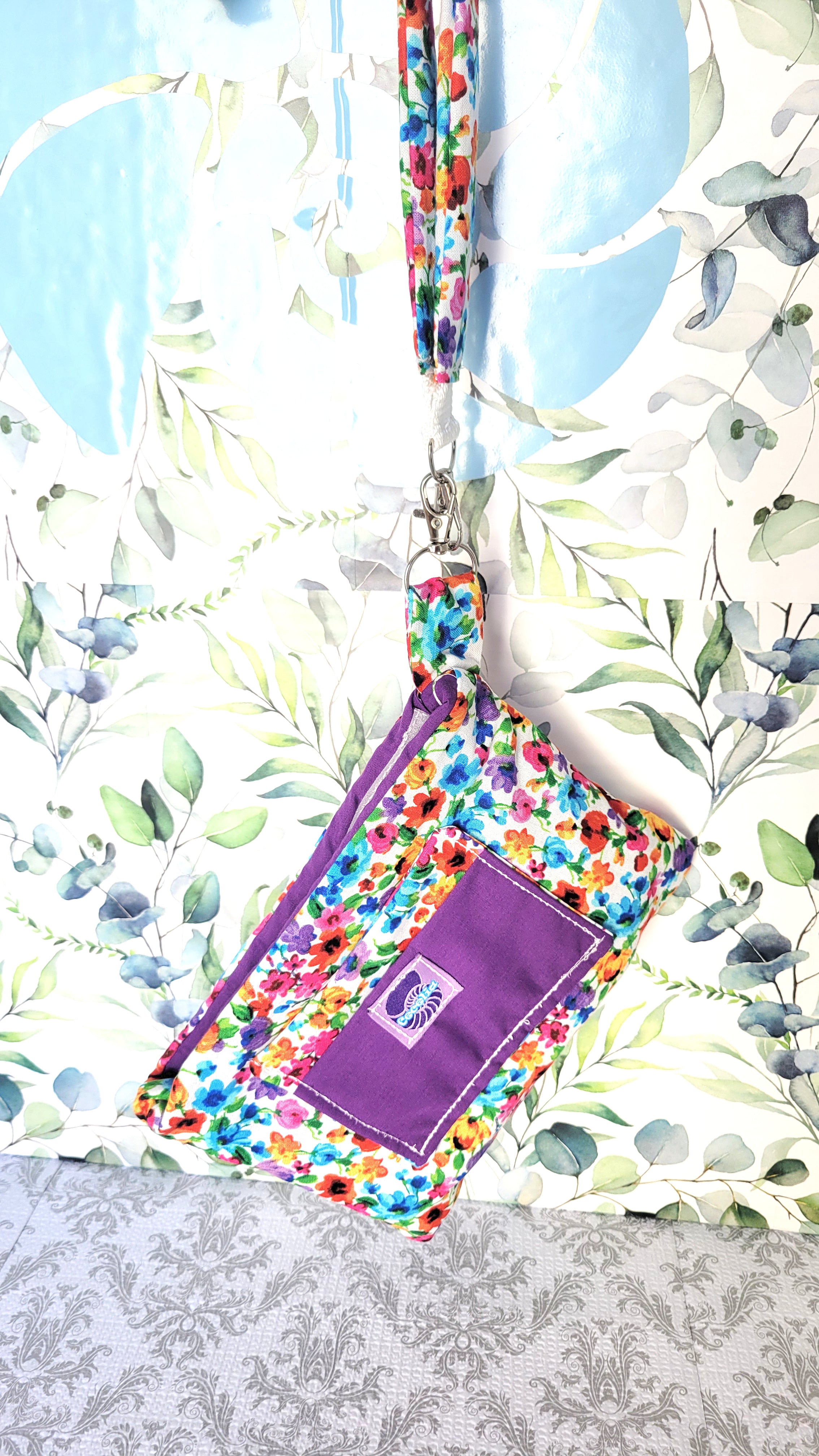 colorful Floral-patterned pouch purse in white with a purple pocket on a textured white background. Hanging on a hook by its hand strap.