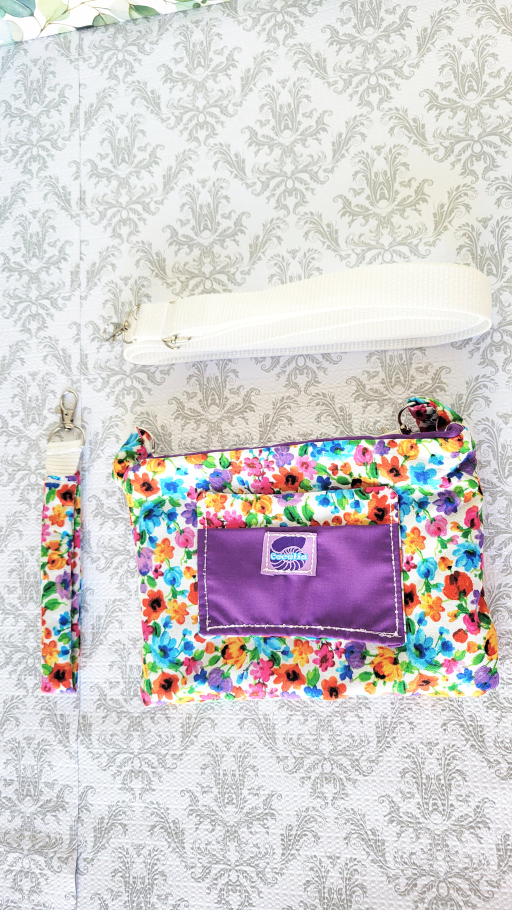 colorful Floral-patterned pouch purse in white with a purple pocket on a textured white background. On top is a white adjustable strap and on the left side of the pouch is a hand strap.