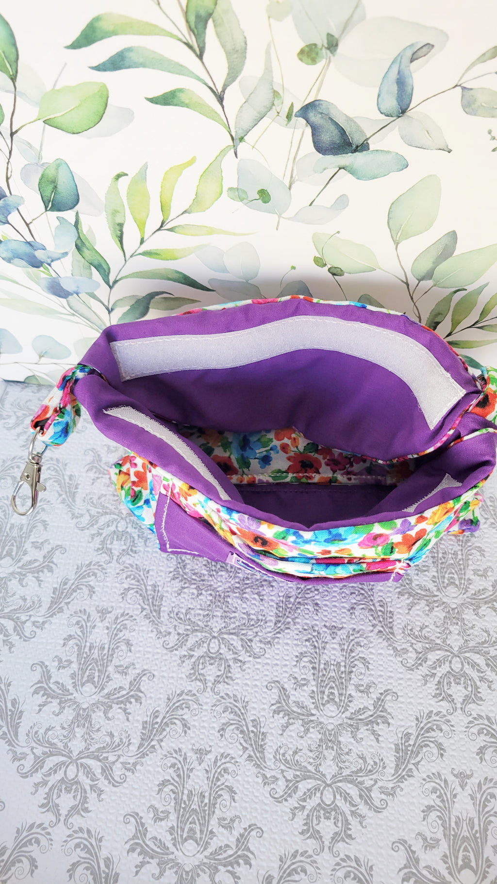 colorful Floral-patterned pouch purse in white with a purple pocket on a textured white background. Open to show a flap with a white velcro as the closure. 