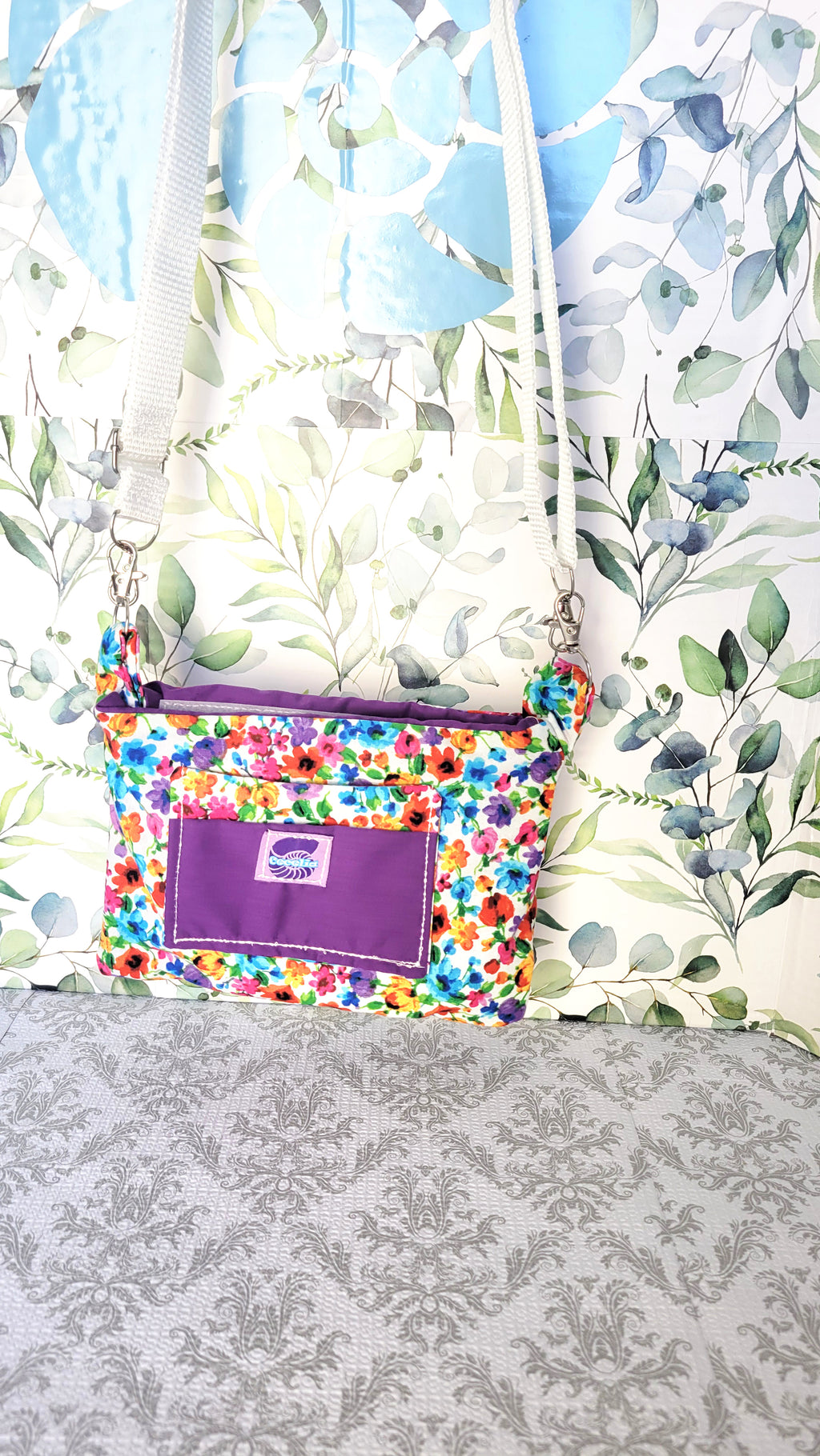 colorful Floral-patterned pouch purse in white with a purple pocket on a textured white background. Hang from its adjustable white strap.