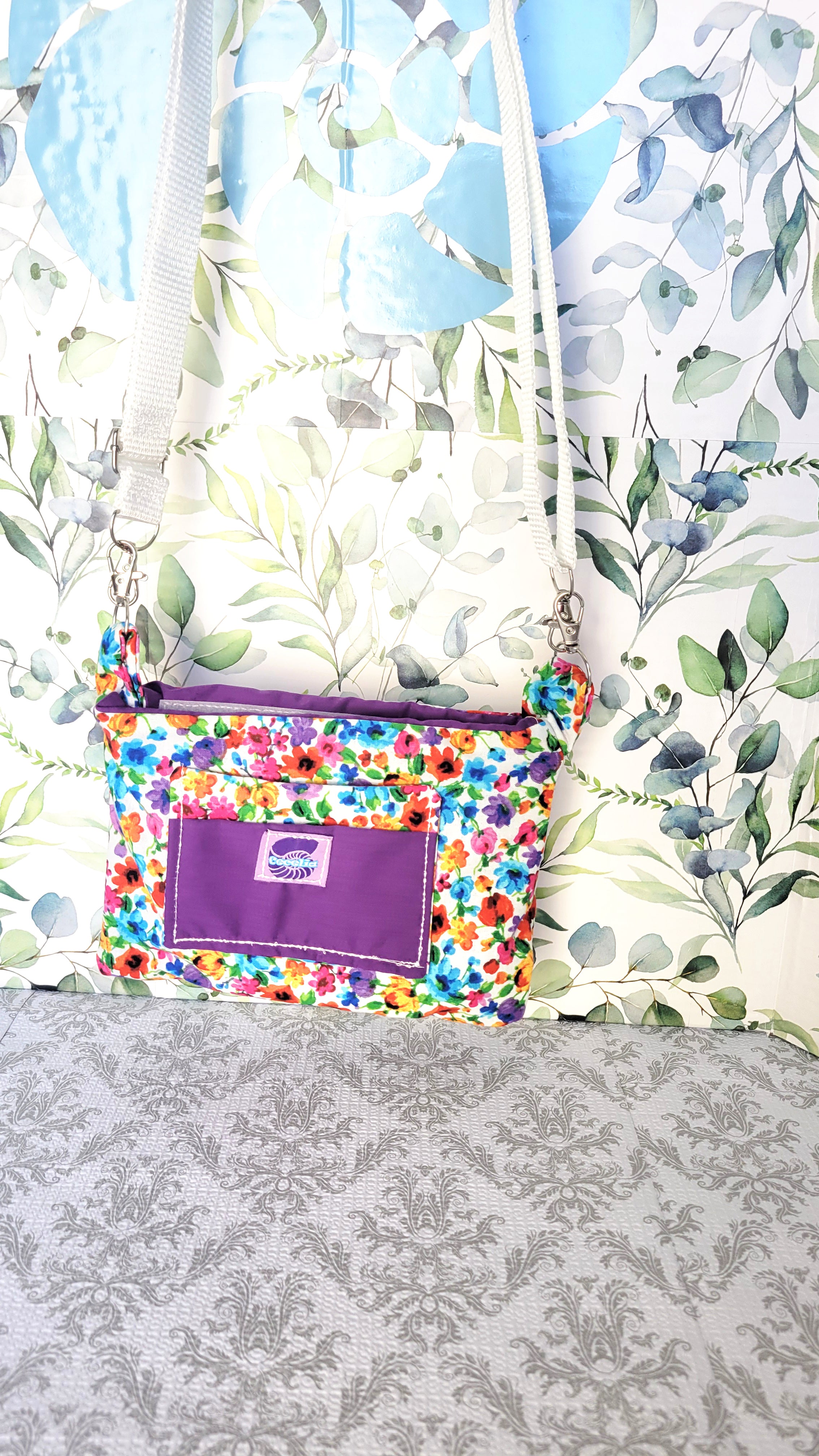 colorful Floral-patterned pouch purse in white with a purple pocket on a textured white background. Hang from its adjustable white strap.