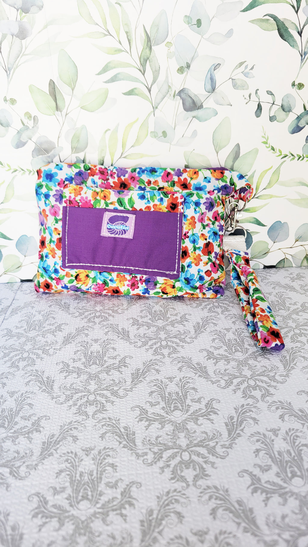 colorful Floral-patterned pouch purse in white with a purple pocket on a textured white background. The pouch has a right side hand strap attached.