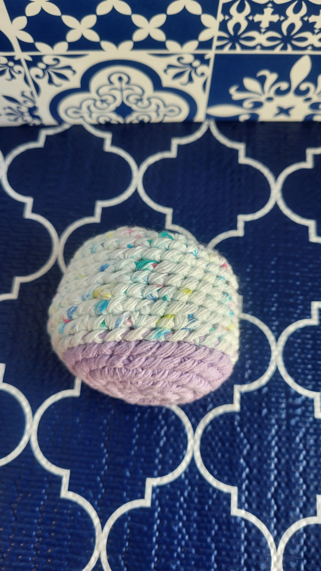 A white with sprinkles and lavender crochet stress ball on a blue patterned fabric background