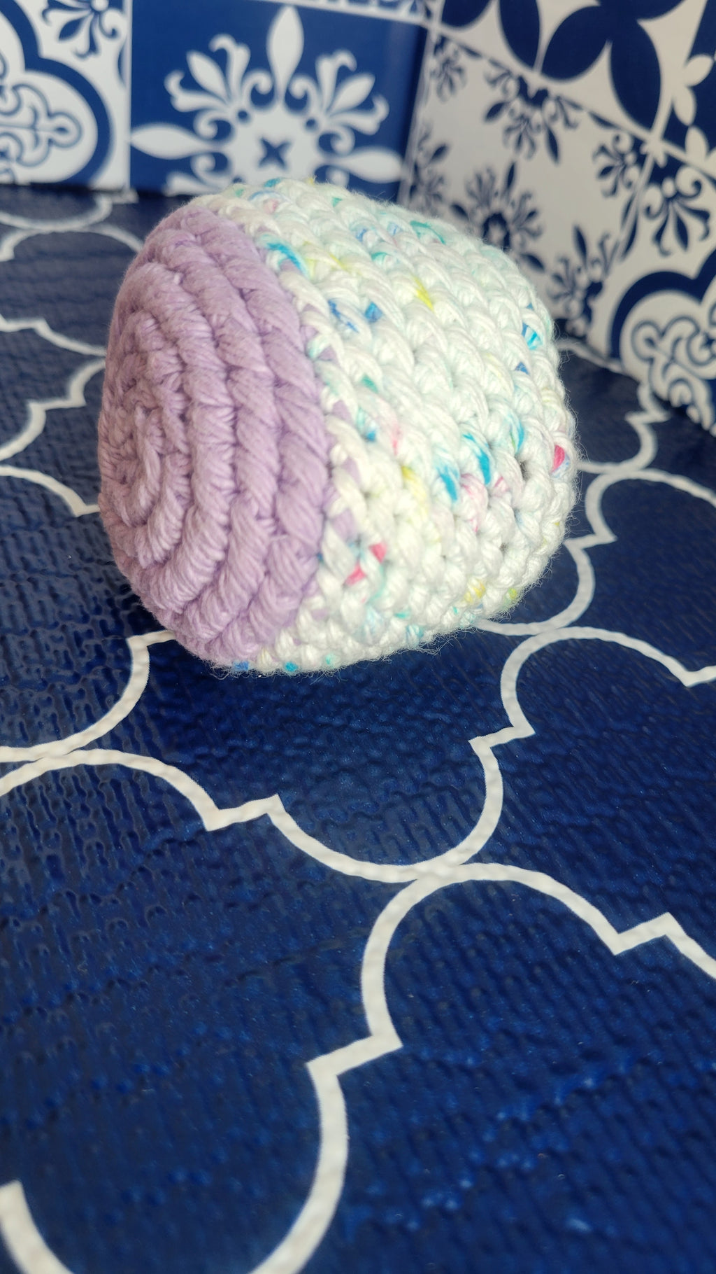 A white with sprinkles and lavender crochet stress ball on a blue patterned fabric background