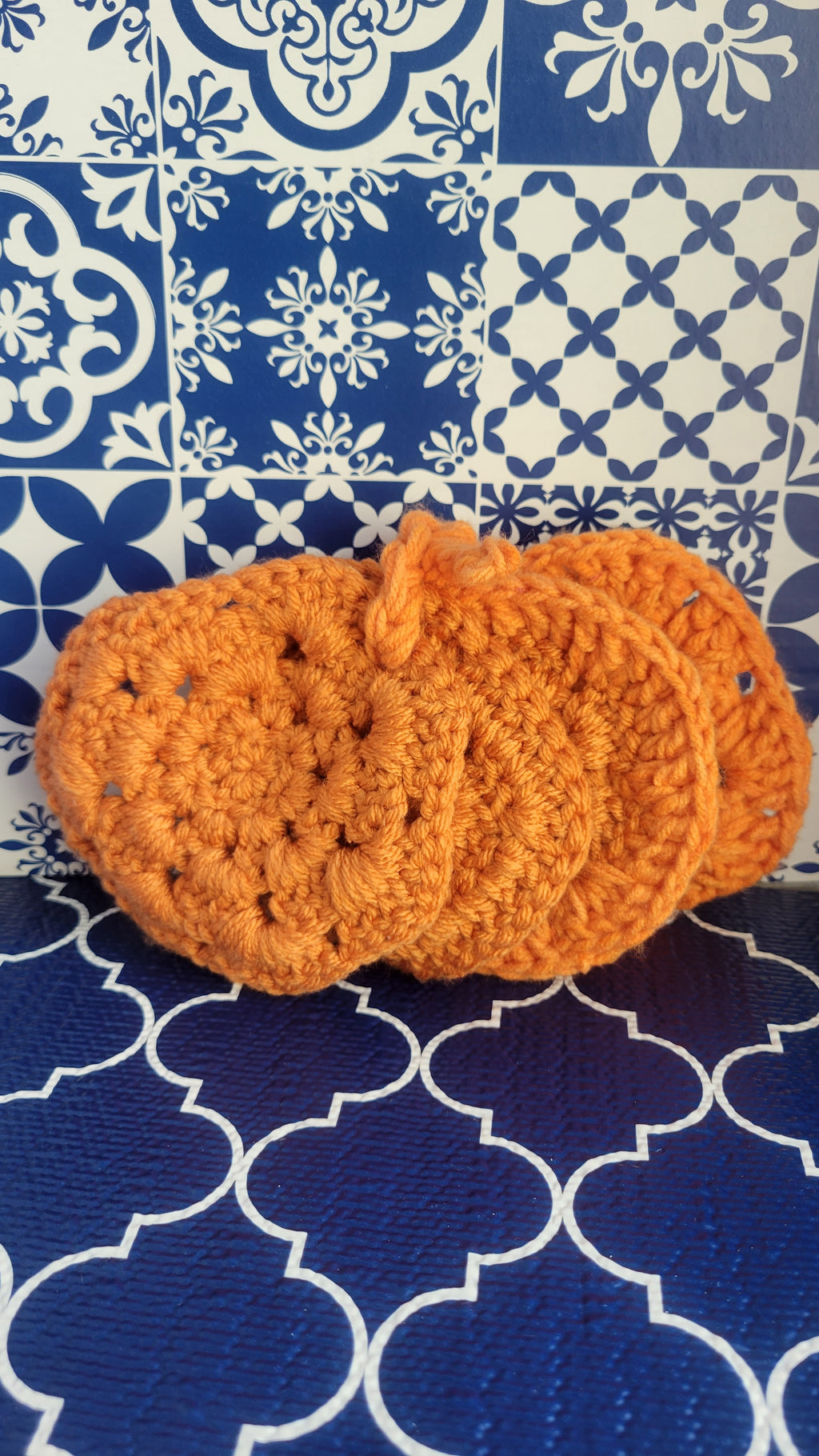 Four orange crocheted coasters in the shape of a pumpkin on a blue and white patterned surface