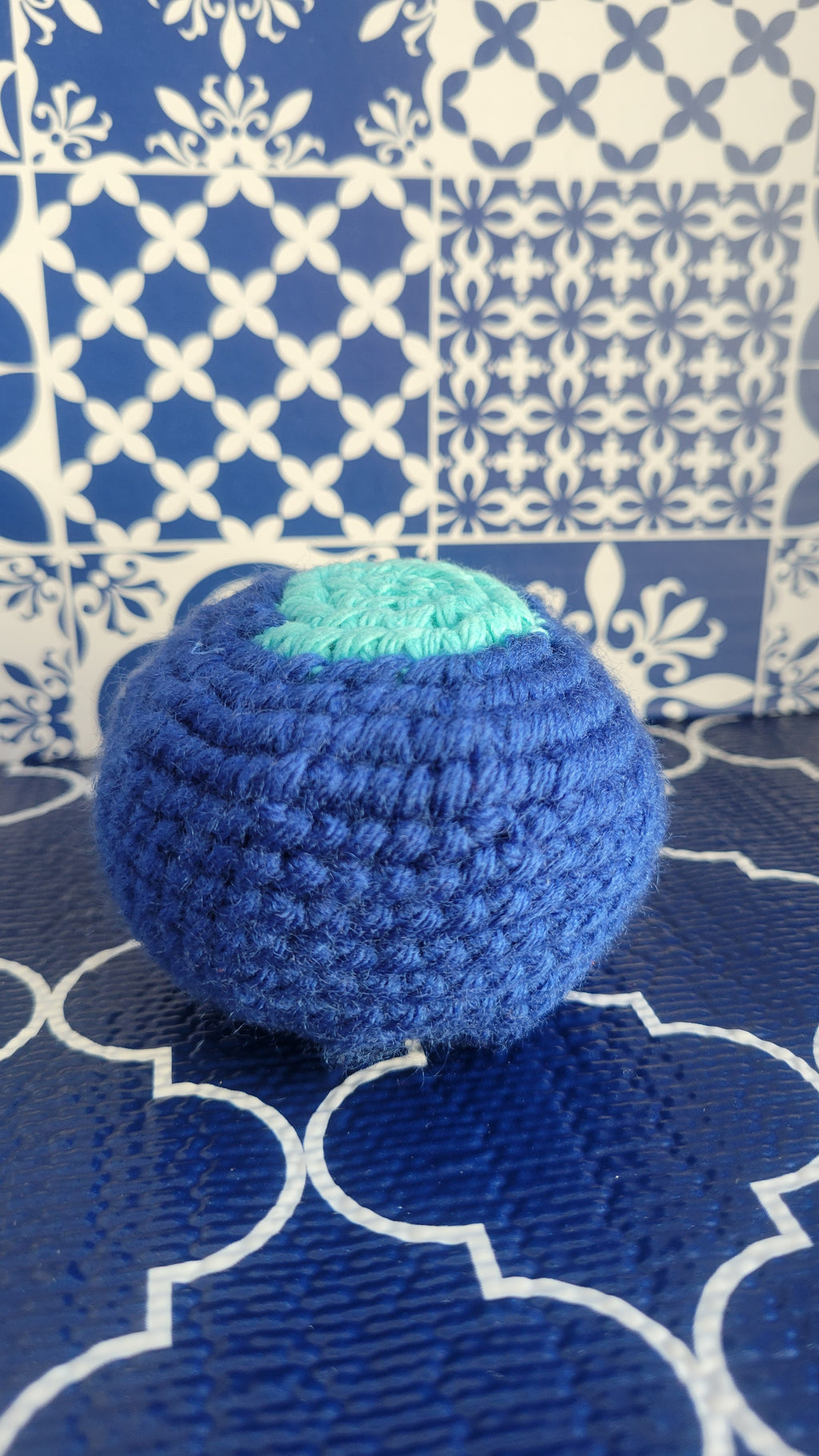 Blue crochet ball with a middle center of light blue on the top. Placed on a blue and white patterned fabric background