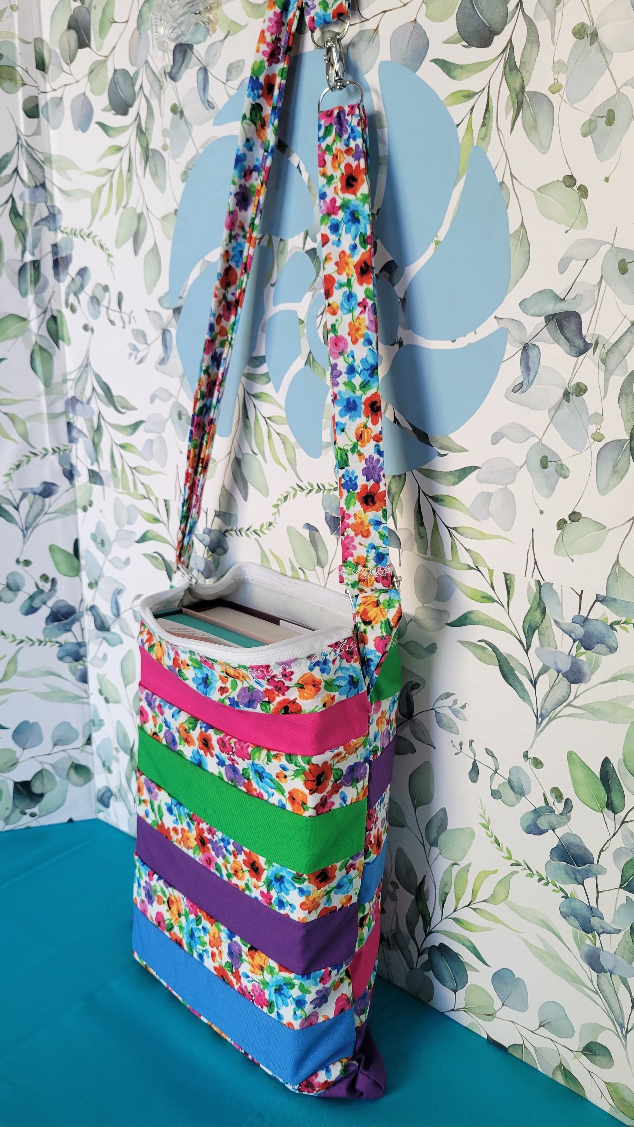 Colorful floral-patterned bag hanging on its adjustable strap in the same pattern as the purse. The purse is made of strips of the colorful floral patterned and in between the strips are strip with the colors of green, purple, blue, and pink. 