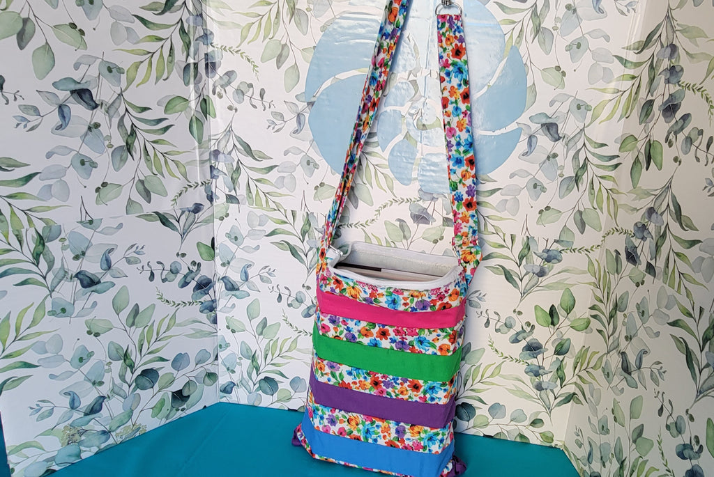 Colorful floral-patterned bag hanging on its adjustable strap in the same pattern as the purse. The purse is made of strips of the colorful floral patterned and in between the strips are strip with the colors of green, purple, blue, and pink. 