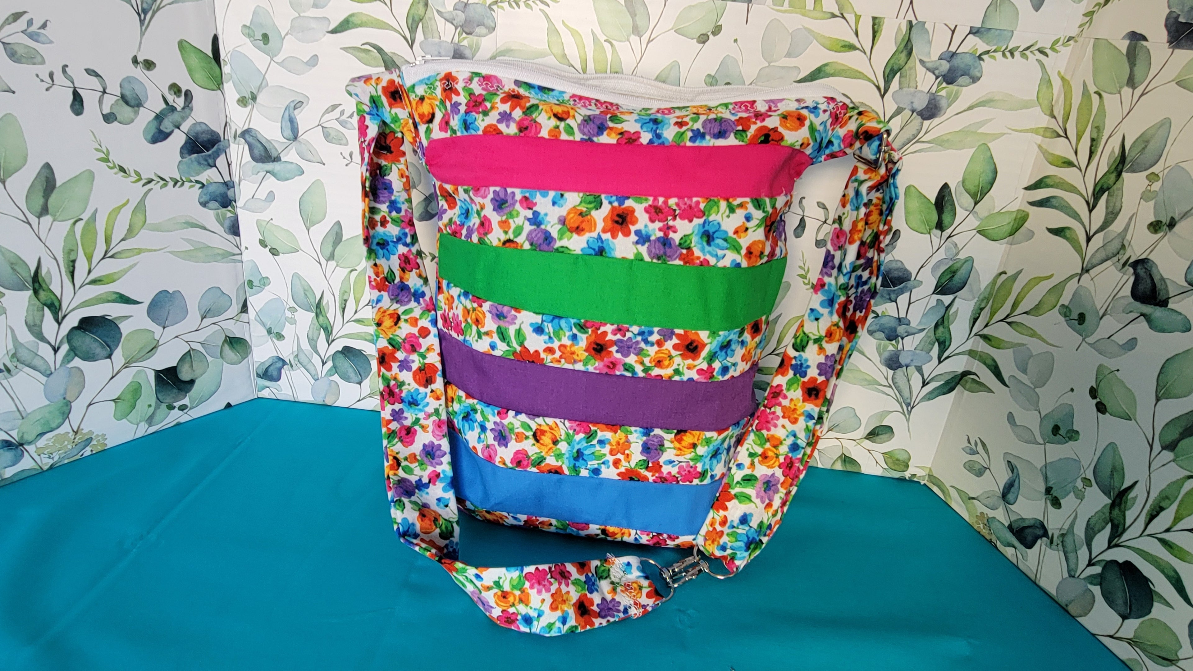 Colorful floral-patterned bag hanging on its adjustable strap in the same pattern as the purse. The purse is made of strips of the colorful floral patterned and in between the strips are strip with the colors of green, purple, blue, and pink. It is closed and standing on its own.