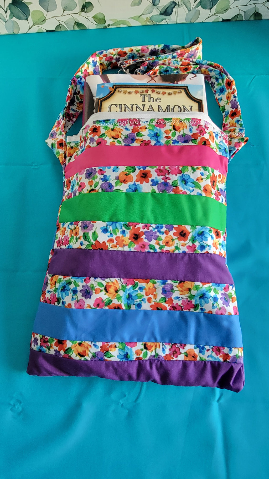 Colorful floral-patterned bag hanging on its adjustable strap in the same pattern as the purse. The purse is made of strips of the colorful floral patterned and in between the strips are strip with the colors of green, purple, blue, and pink. It is open to show its one pocket inside the purse. The inside is all white.