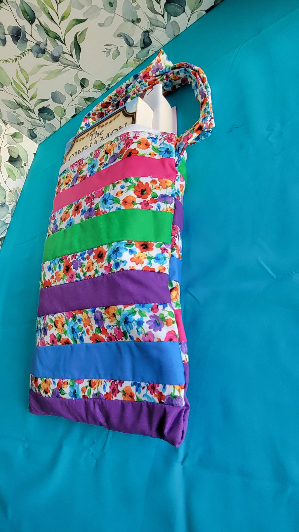 Colorful floral-patterned bag hanging on its adjustable strap in the same pattern as the purse. The purse is made of strips of the colorful floral patterned and in between the strips are strip with the colors of green, purple, blue, and pink. 