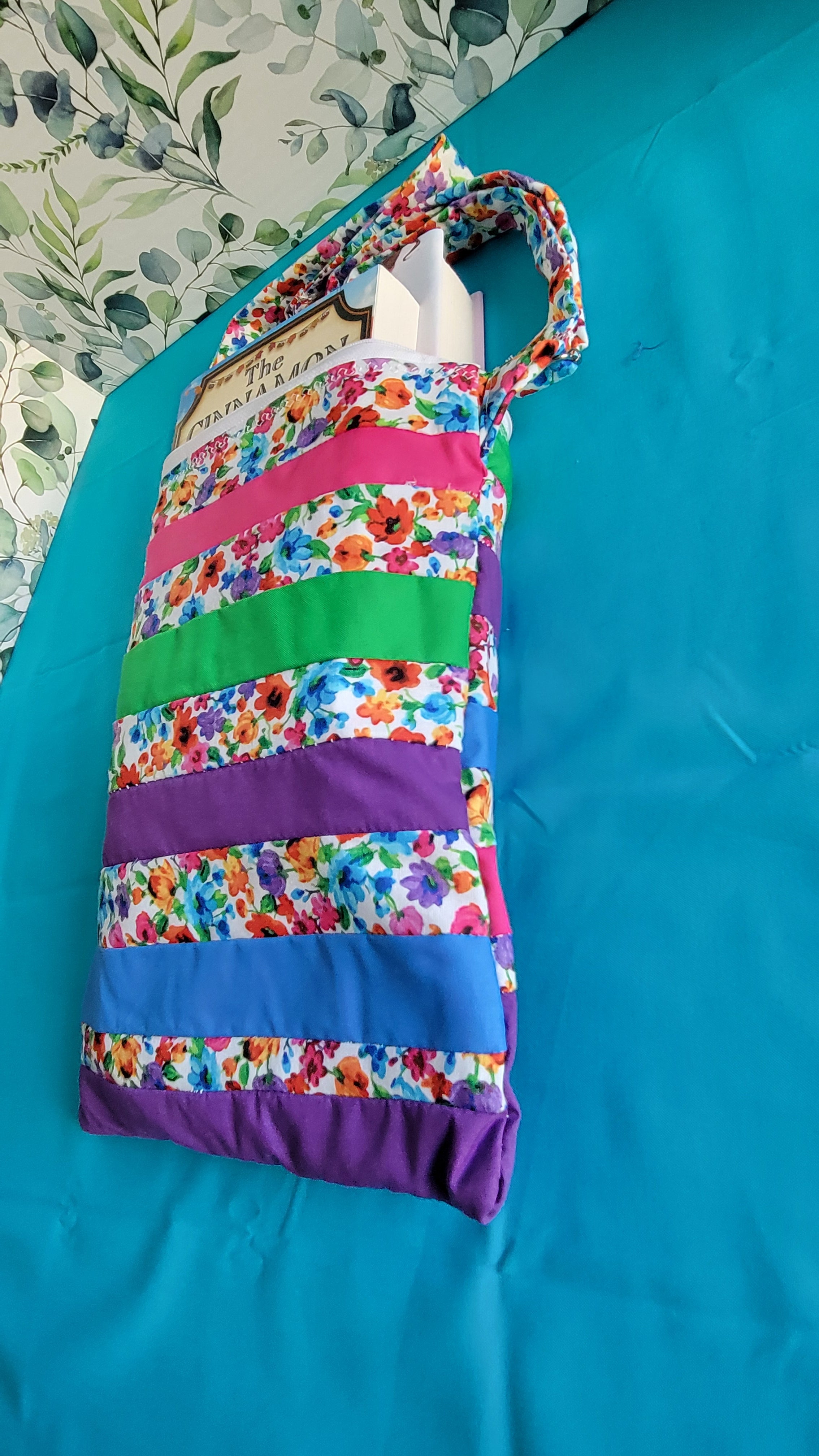 Colorful floral-patterned bag hanging on its adjustable strap in the same pattern as the purse. The purse is made of strips of the colorful floral patterned and in between the strips are strip with the colors of green, purple, blue, and pink. 