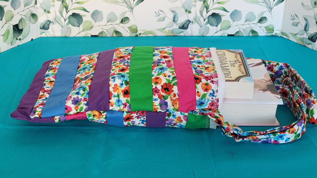 Colorful floral-patterned bag hanging on its adjustable strap in the same pattern as the purse. The purse is made of strips of the colorful floral patterned and in between the strips are strip with the colors of green, purple, blue, and pink. It is open to show its one pocket inside the purse. The inside is all white.