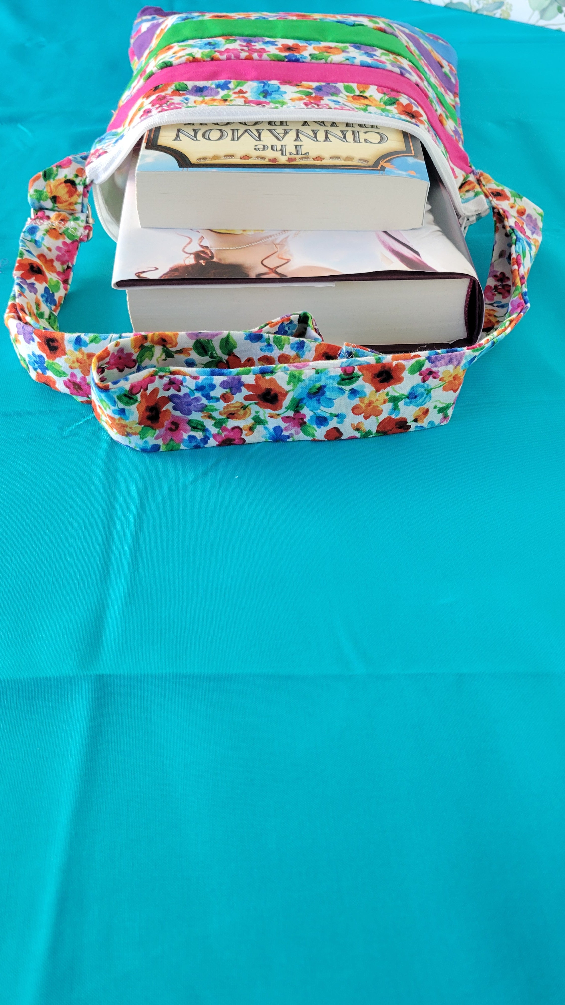 Colorful floral-patterned bag hanging on its adjustable strap in the same pattern as the purse. The purse is made of strips of the colorful floral patterned and in between the strips are strip with the colors of green, purple, blue, and pink. It is open to show its one pocket inside the purse. The inside is all white.