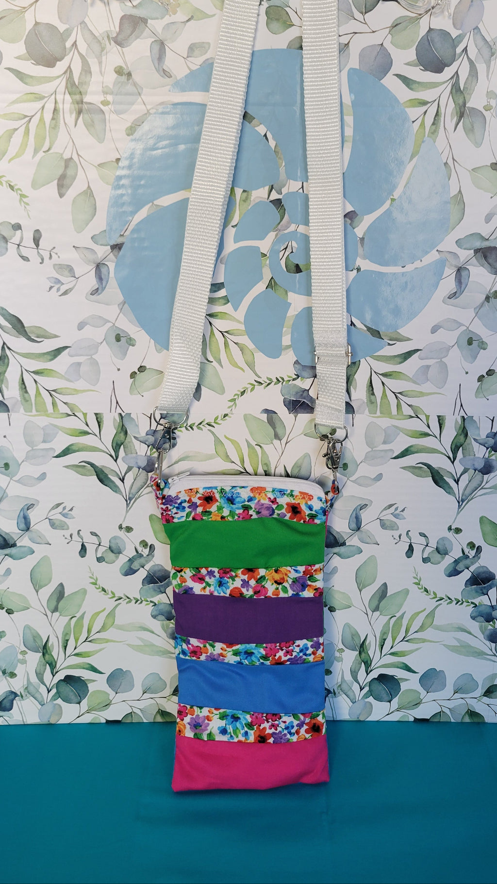 Colorful phone purse with a floral pattern. The pattern is in strips and in between the strip colors of green, purple, blue, and pink. The purse is hanging from its adjustable white strap.