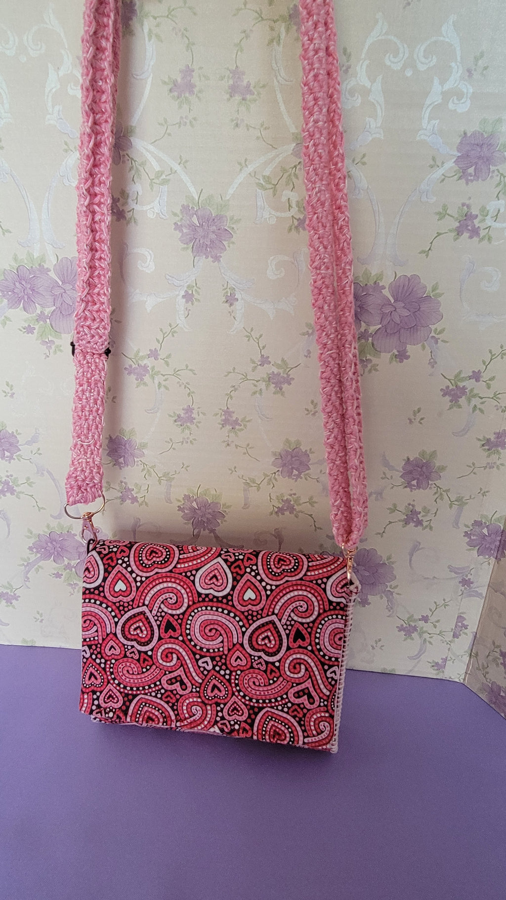 Pink Flores Clutch Purse with a ripple effect of hearts in a selection of colors pink, white, red and black. Hanging in front of a tan wallpaper with purple colors.