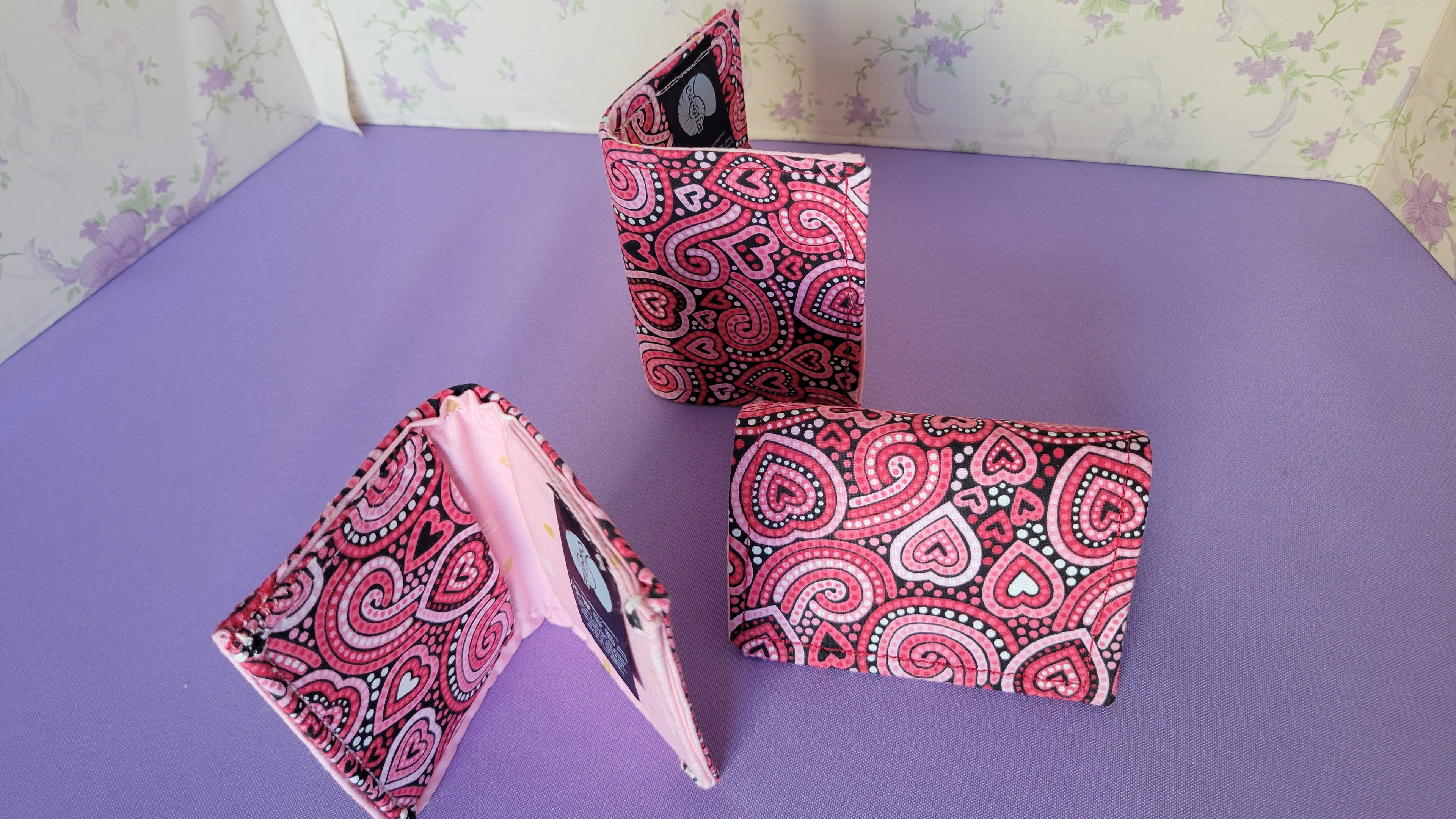 Three Javier wallets with a ripple effect of hearts in a selection of colors pink, white, red and black. They are placed parallel with one on a turning table.