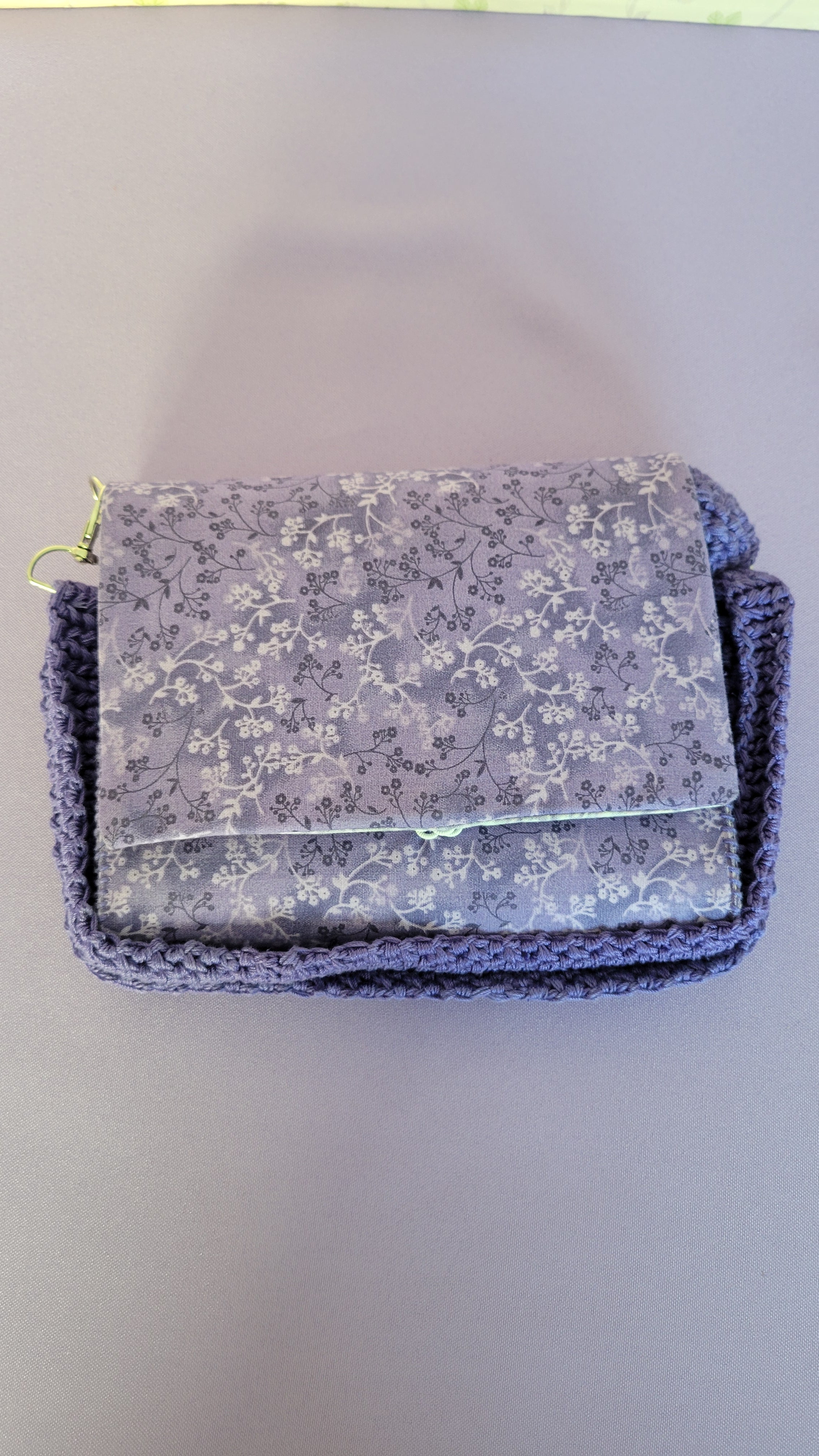 Purple clutch flores purse bag with floral patterns on a purple fabric. Around the purse is a crochet adjustable strap.