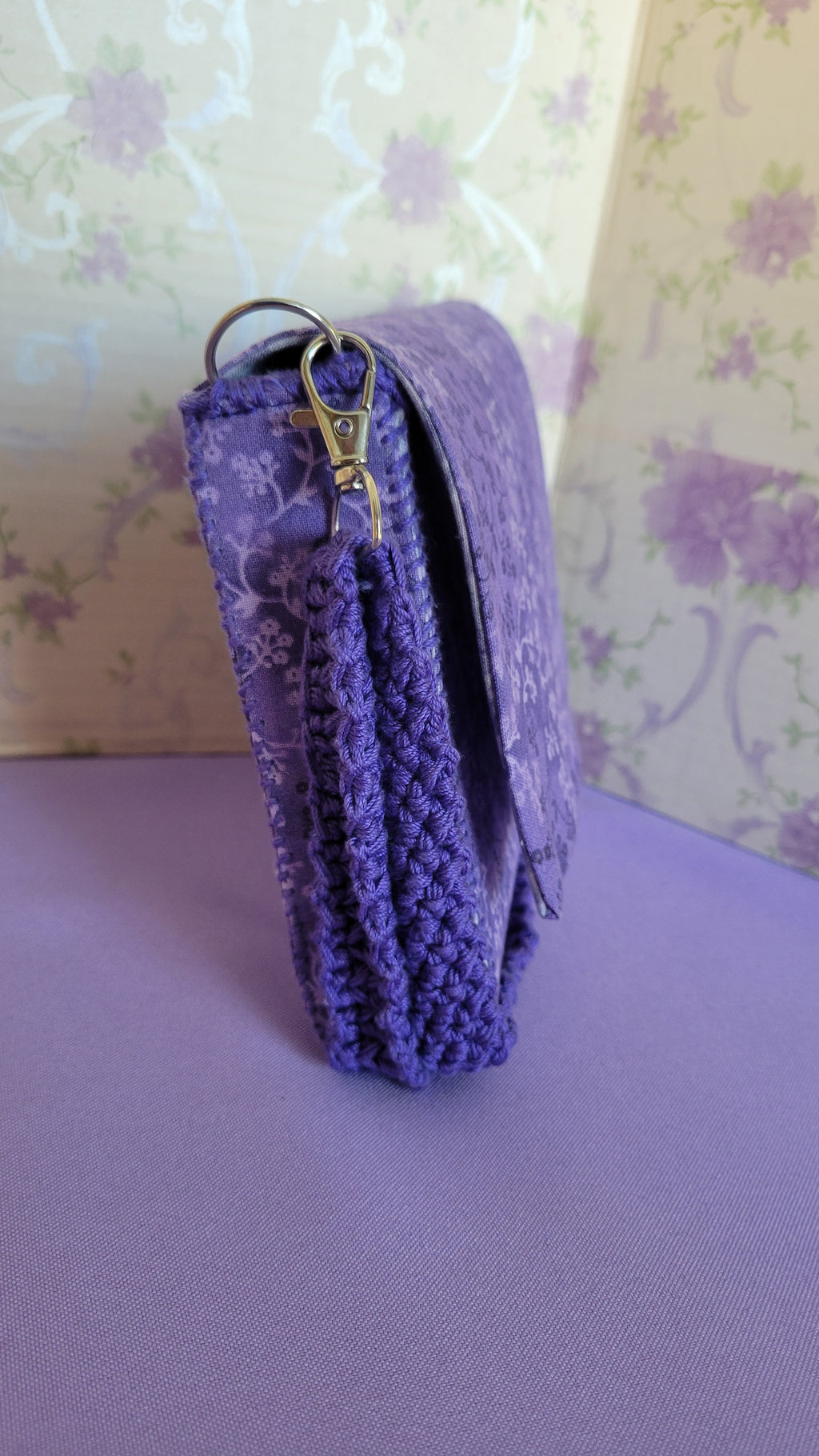 Side profiles of the purple clutch flores purse bag with floral patterns on a purple fabric. The side profiles shows the crochet  adjustable strap in a color similar to the purse color.