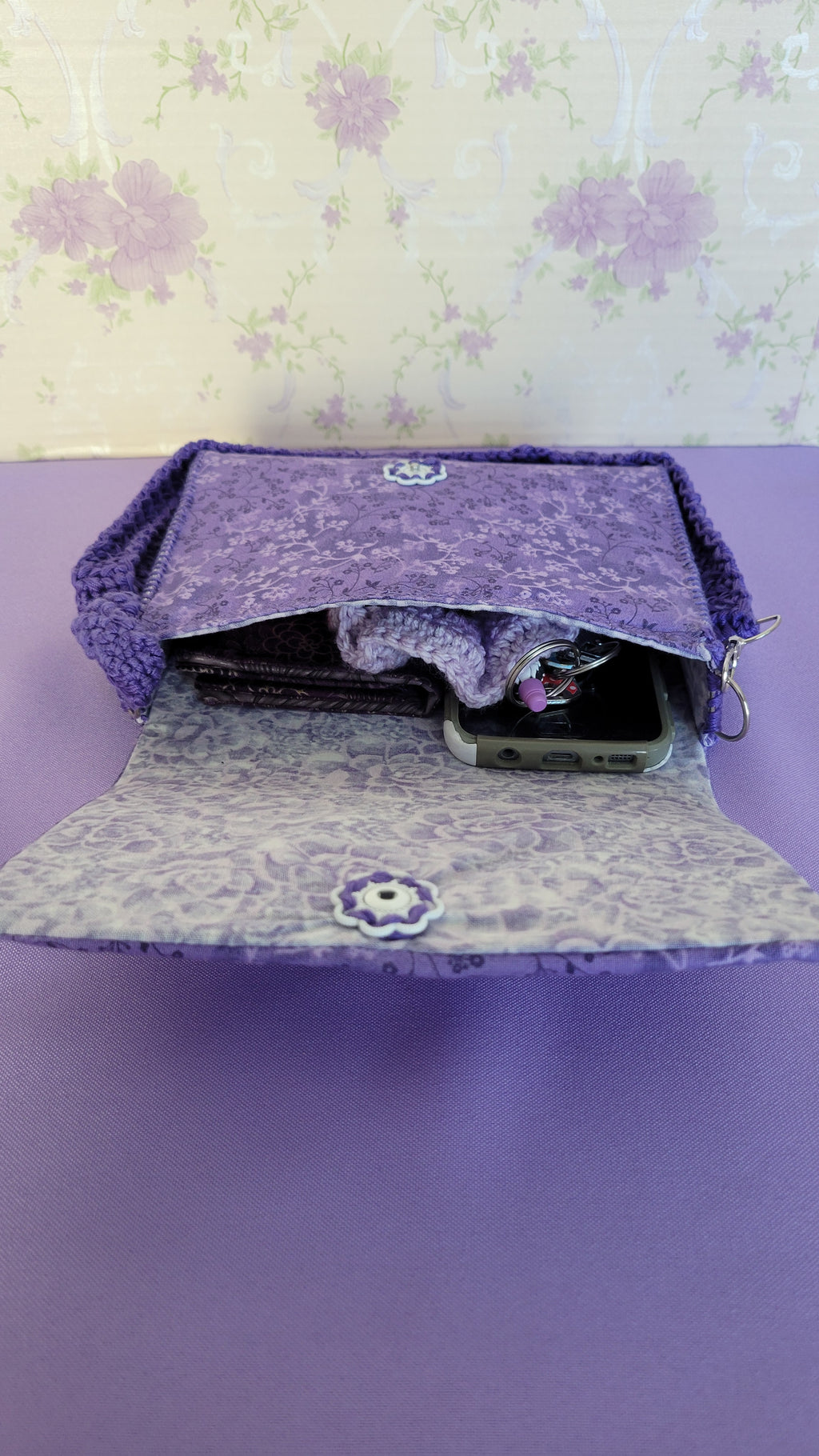Purple clutch flores purse bag with floral patterns on a purple fabric. Inside shows all the items it can fit. A wallet, car keys, scrunchies, and more.