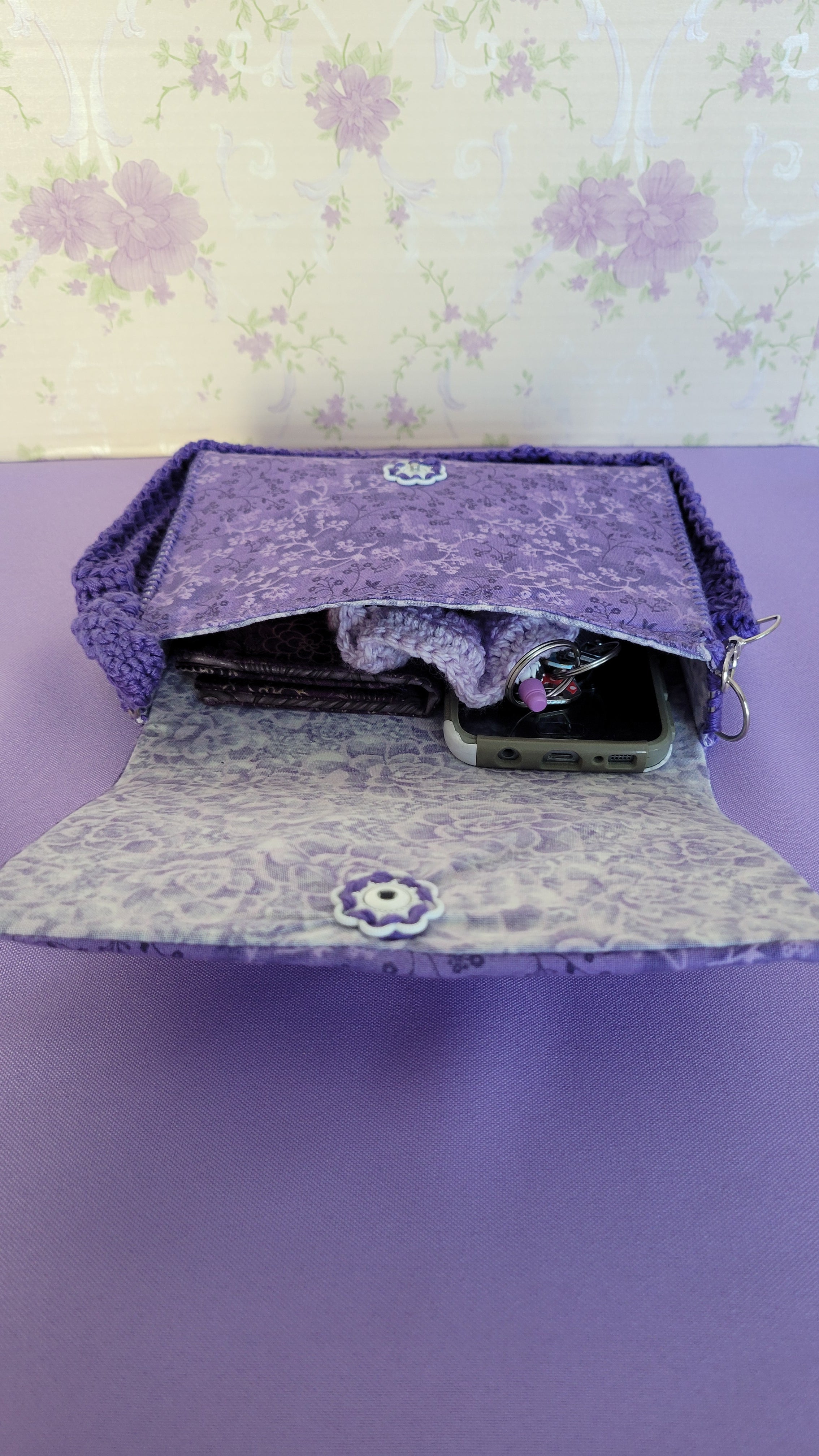 Purple clutch flores purse bag with floral patterns on a purple fabric. Inside shows all the items it can fit. A wallet, car keys, scrunchies, and more.