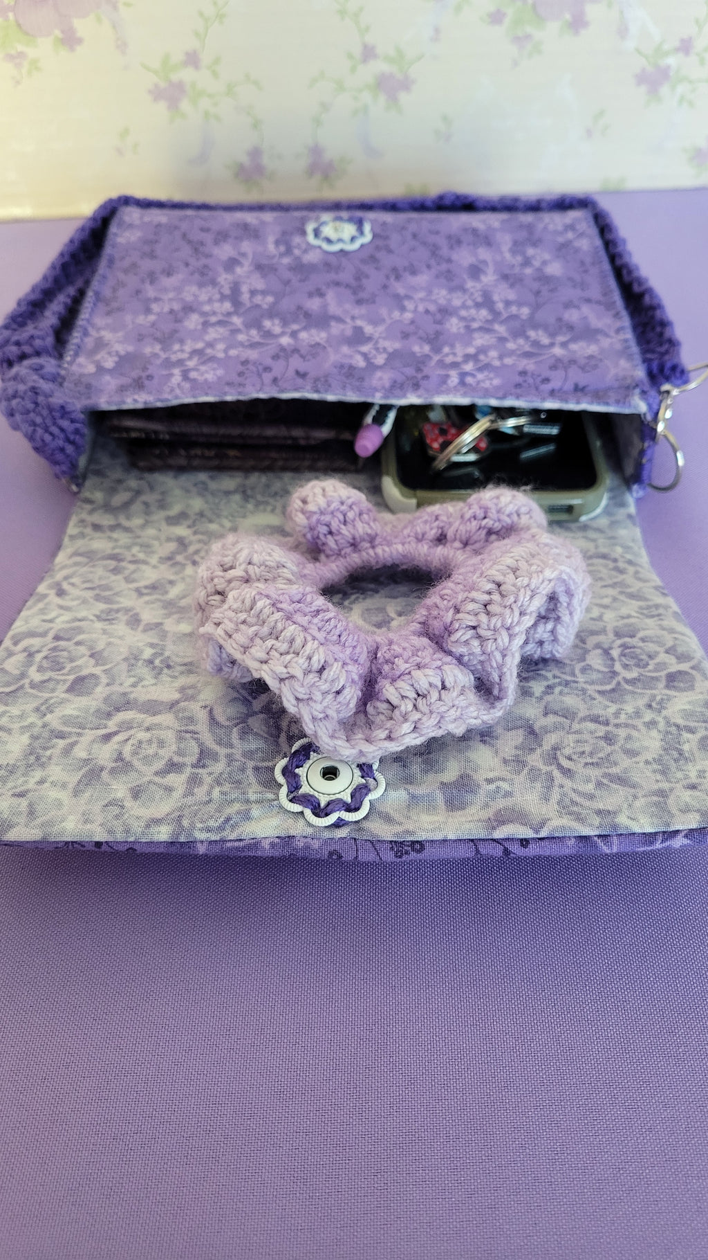 Purple clutch flores purse bag with floral patterns on a purple fabric. The purse is open showing a lavender design of roses, and a white vintage flower push bottom.
Inside of the purse shows all the items it can fit. A wallet, car keys, scrunchies, and more.