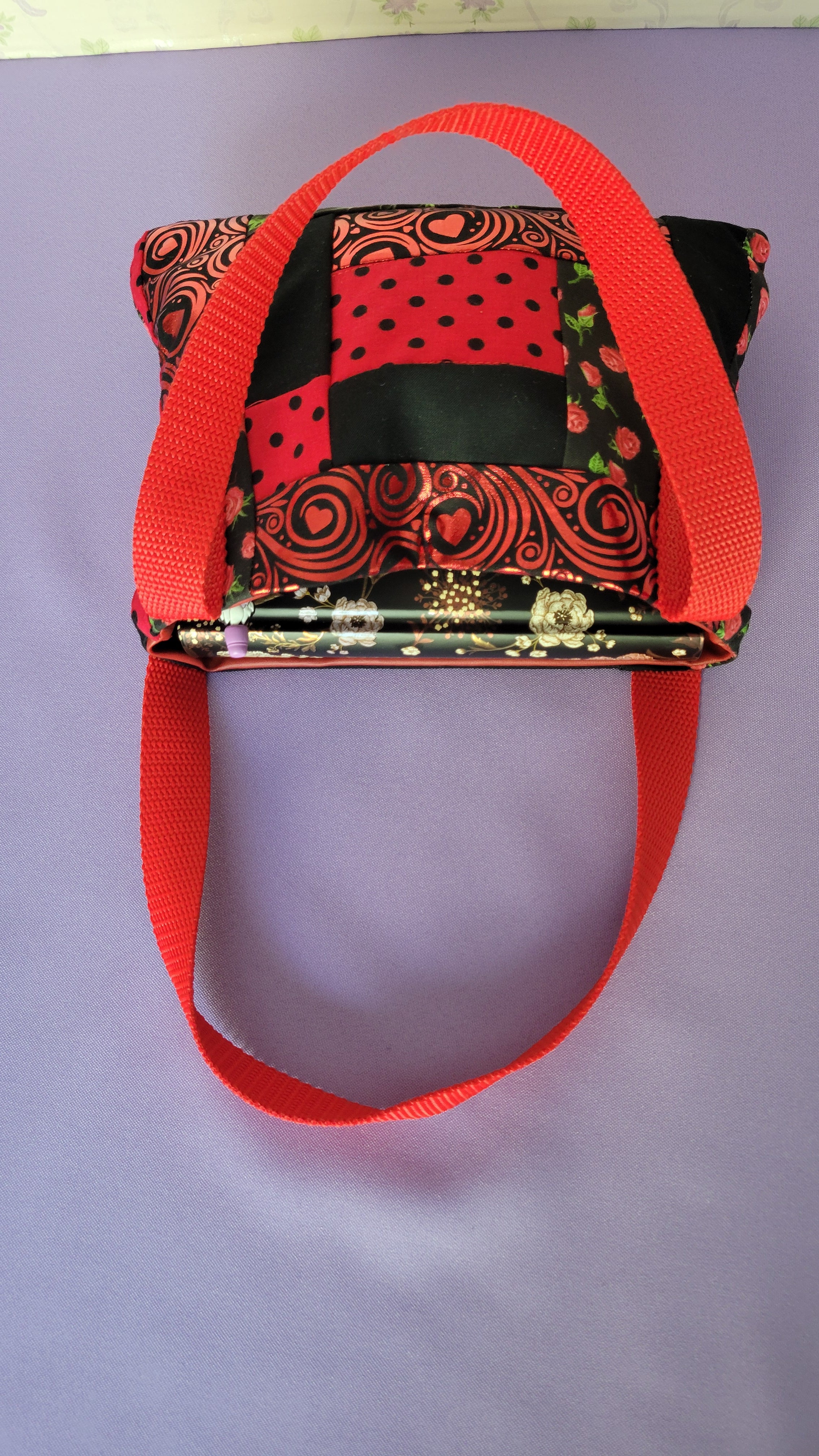 A Red Toe bag called Tracy style with two straps in red. The 5 quilt style bag has the color black., red with black polka dots, red swirls and hearts in black background, and red roses with green leaves pattern on background fabric. 

Inside of the purse is items it can fit. A hand sanitizer, a red heart wallet, an example book it can fit which is emma, a touch phone and Ursula pen.