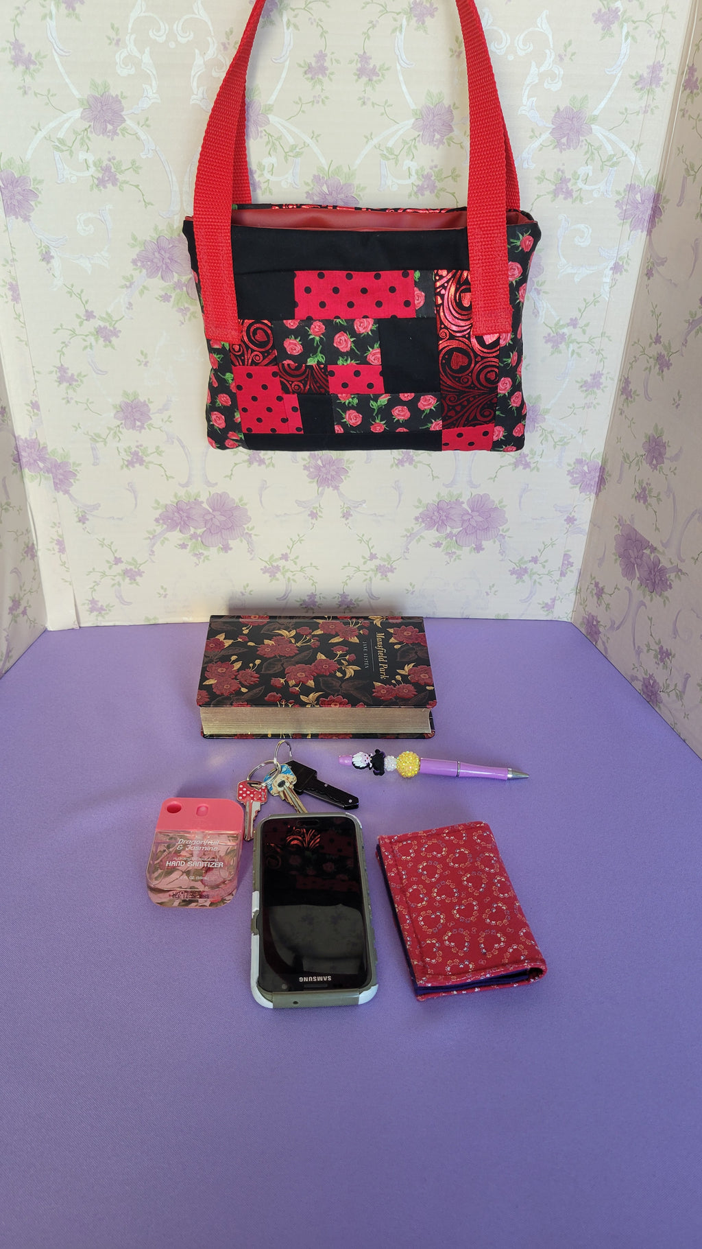 A Red Toe bag called Tracy style with two straps in red. The 5 quilt style bag has the color black., red with black polka dots, red swirls and hearts in black background, and red roses with green leaves pattern on background fabric. 

In front of the purse is items it can fit. A hand sanitizer, a red heart wallet, an example book it can fit, a touch phone and Ursula pen.