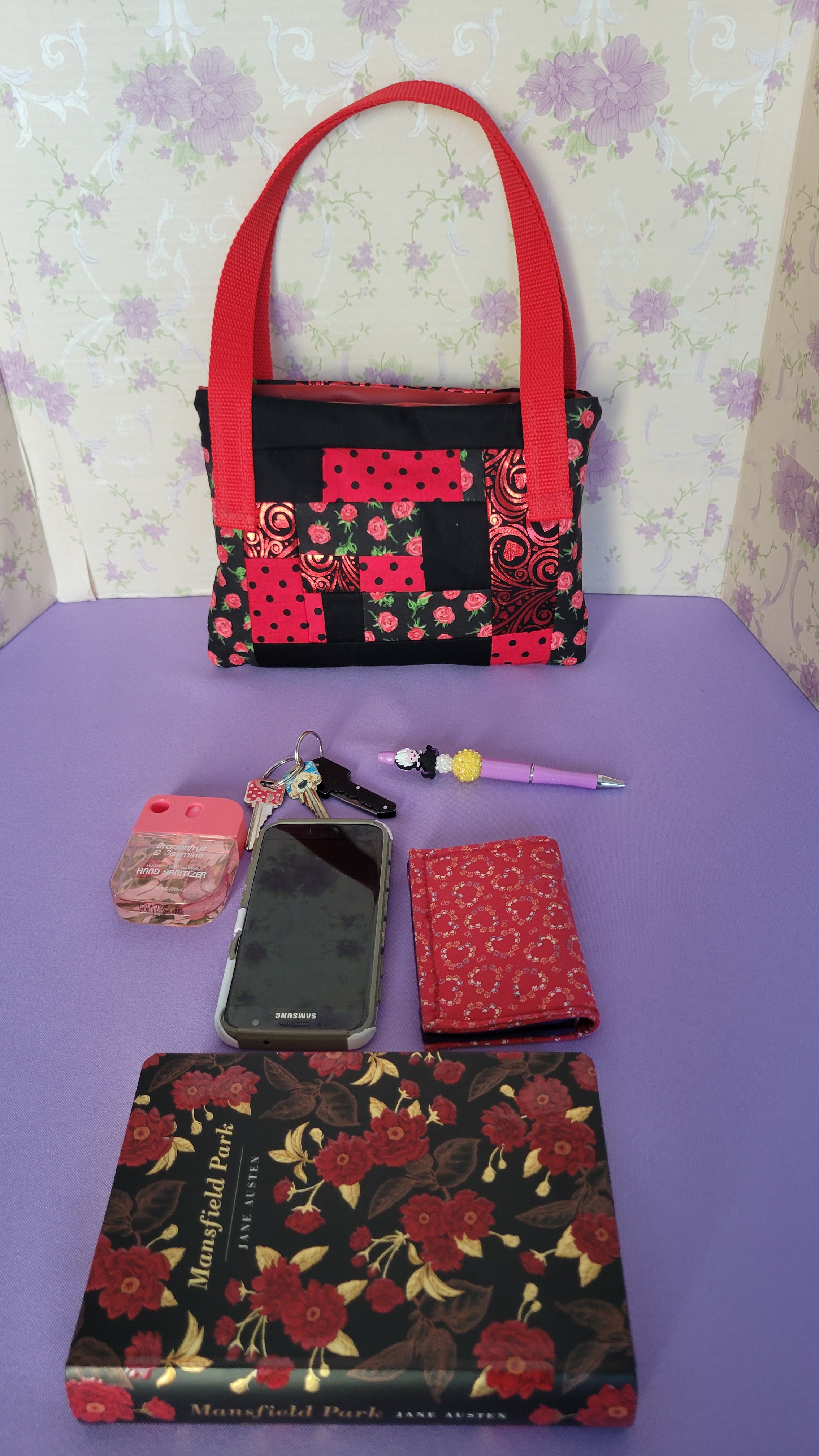 A Red Toe bag called Tracy style with two straps in red. The 5 quilt style bag has the color black., red with black polka dots, red swirls and hearts in black background, and red roses with green leaves pattern on background fabric. 

In front of the purse is items it can fit. A hand sanitizer, a red heart wallet, an example book it can fit, a touch phone and Ursula pen.