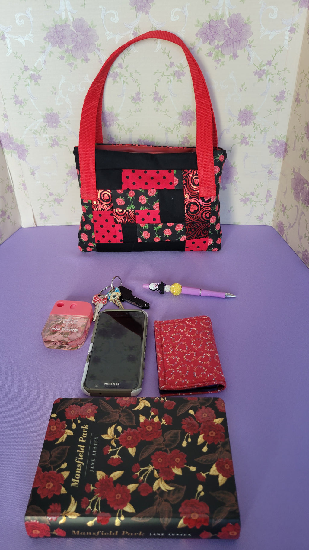 A Red Toe bag called Tracy style with two straps in red. The 5 quilt style bag has the color black., red with black polka dots, red swirls and hearts in black background, and red roses with green leaves pattern on background fabric. 

In front of the purse is items it can fit. A hand sanitizer, a red heart wallet, an example book it can fit, a touch phone and Ursula pen.