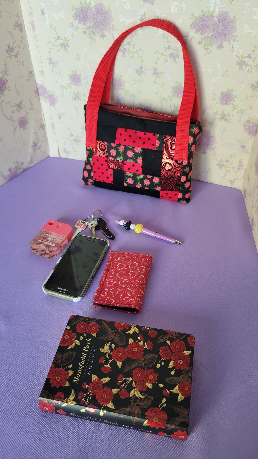 A Red Toe bag called Tracy style with two straps in red. The 5 quilt style bag has the color black., red with black polka dots, red swirls and hearts in black background, and red roses with green leaves pattern on background fabric. 

In  side view of the purse is items it can fit. A hand sanitizer, a red heart wallet, an example book it can fit, a touch phone and Ursula pen.