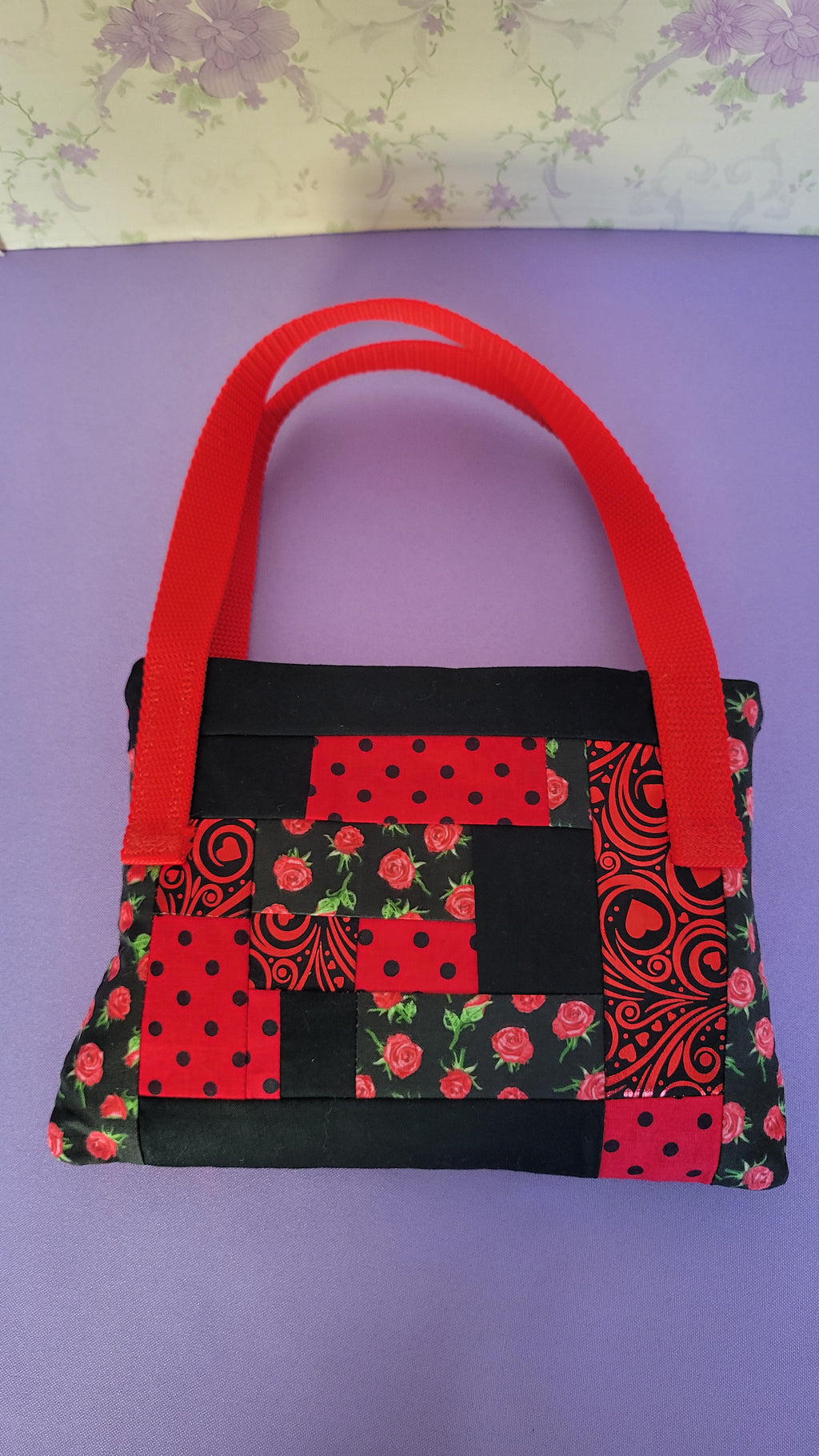 A Red Toe bag called Tracy style with two straps in red. The 5 quilt style bag has the color black., red with black polka dots, red swirls and hearts in black background, and red roses with green leaves pattern on background fabric. 