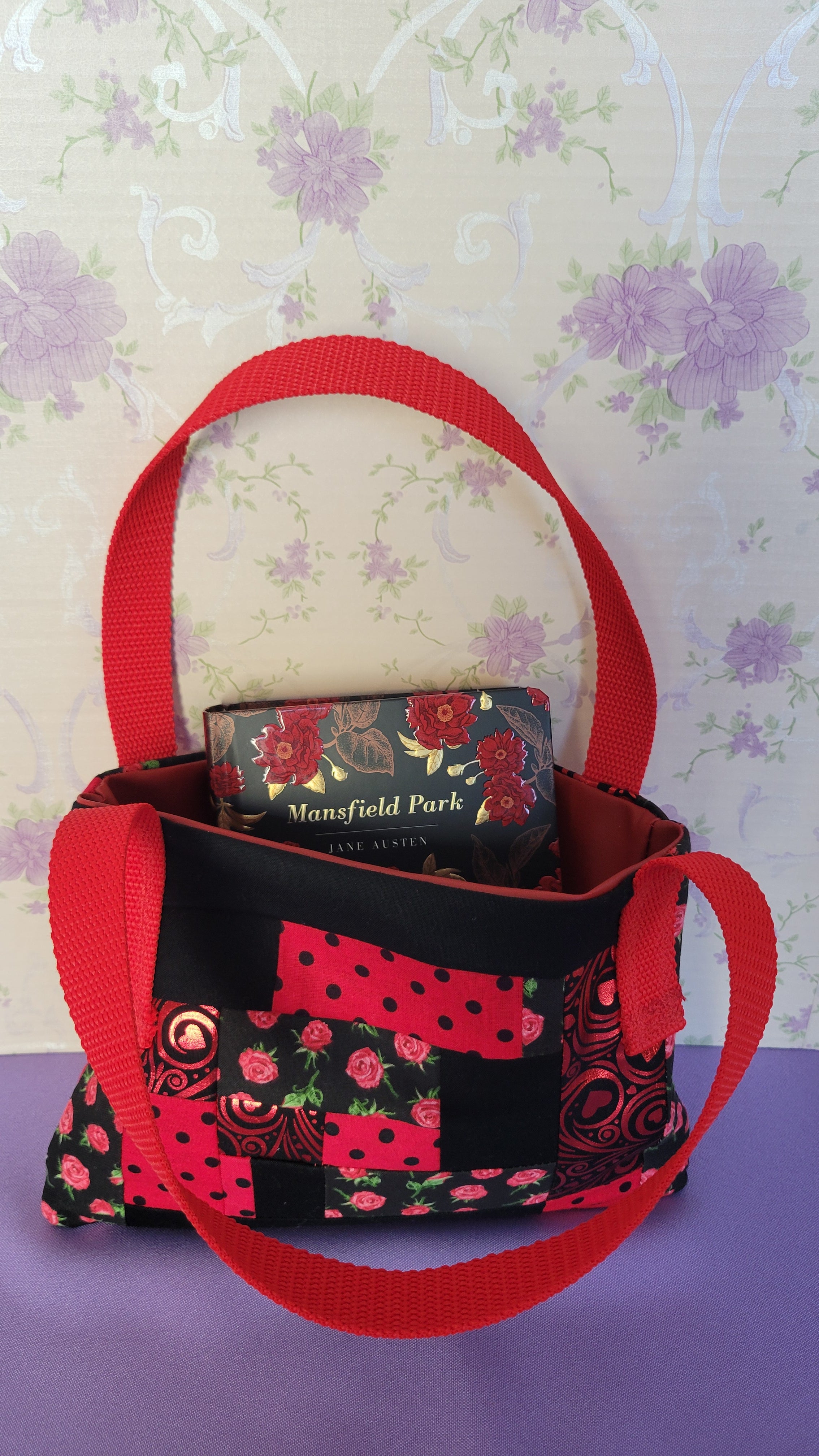 A Red Toe bag called Tracy style with two straps in red. The 5 quilt style bag has the color black., red with black polka dots, red swirls and hearts in black background, and red roses with green leaves pattern on background fabric. 

Inside of the purse is an example book of Manfeild Park.