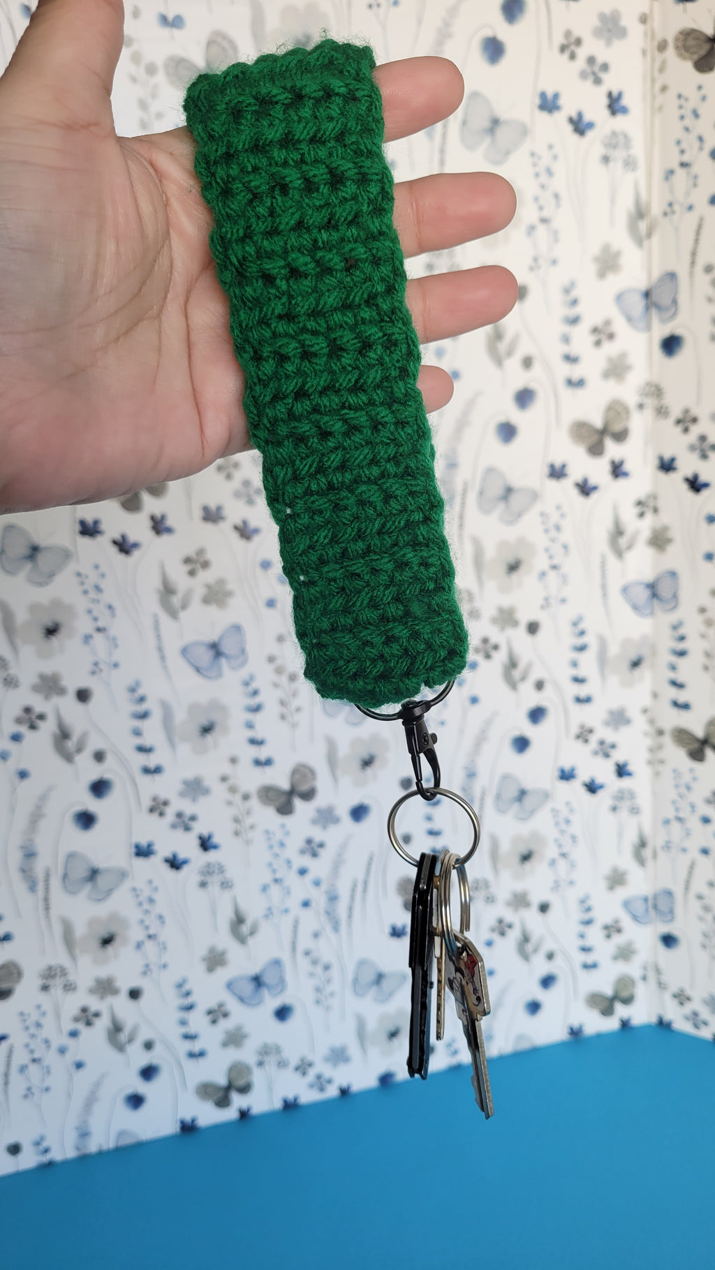 Green crocheted keychain held by a hand on a patterned fabric background. Several key hanging on a black metal hook.