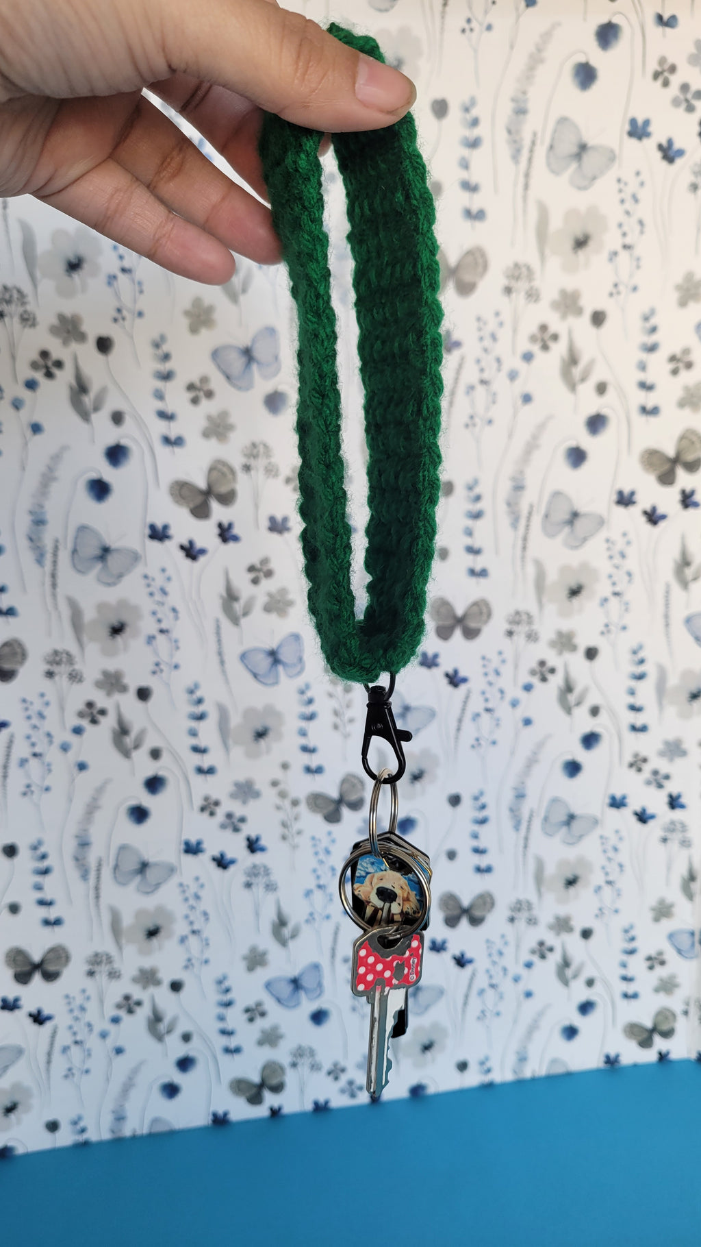 Green crochet bracelet on a wrist with a blue background. Several key hanging on a black metal hook.