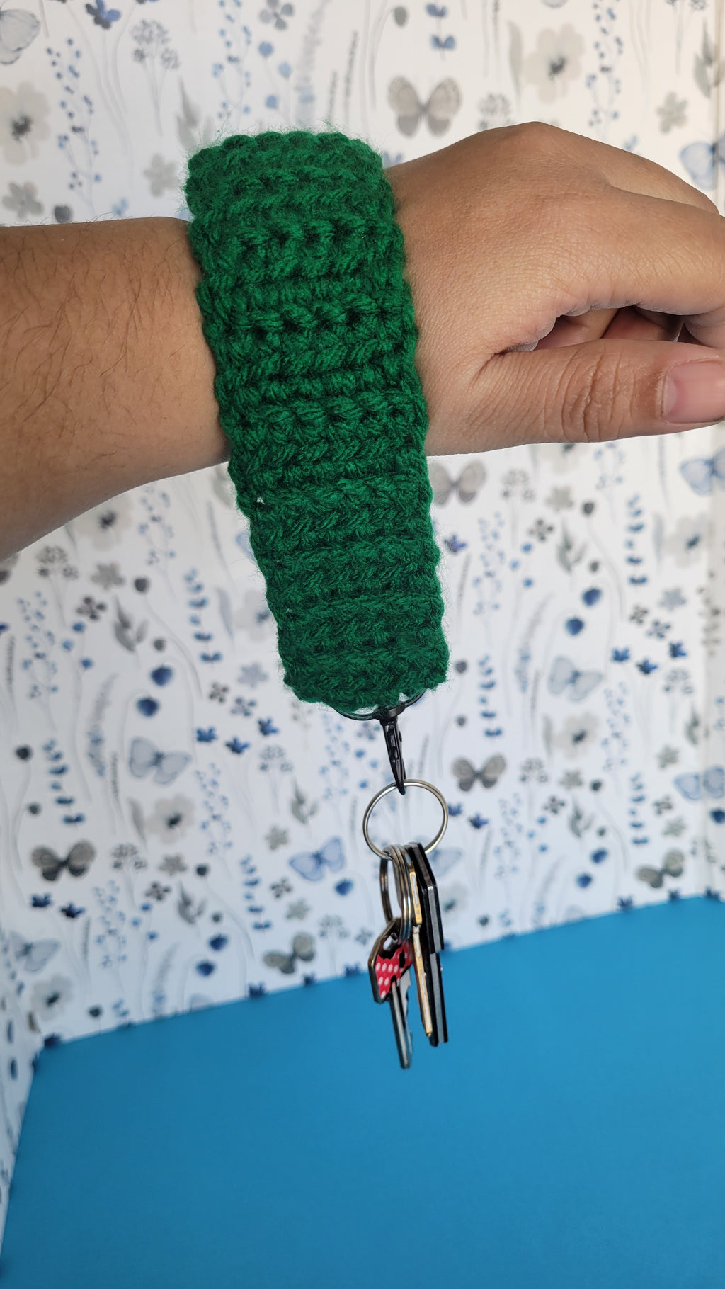 Green crochet bracelet on a wrist with a blue background. Several key hanging on a black metal hook.