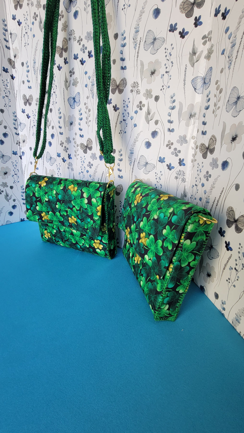 Two green leaf clover fabric clutch purses. One is hanging from it's green lace strap and the second is leaning against a floral patterns on a blue background. Picture is taken from the right angle