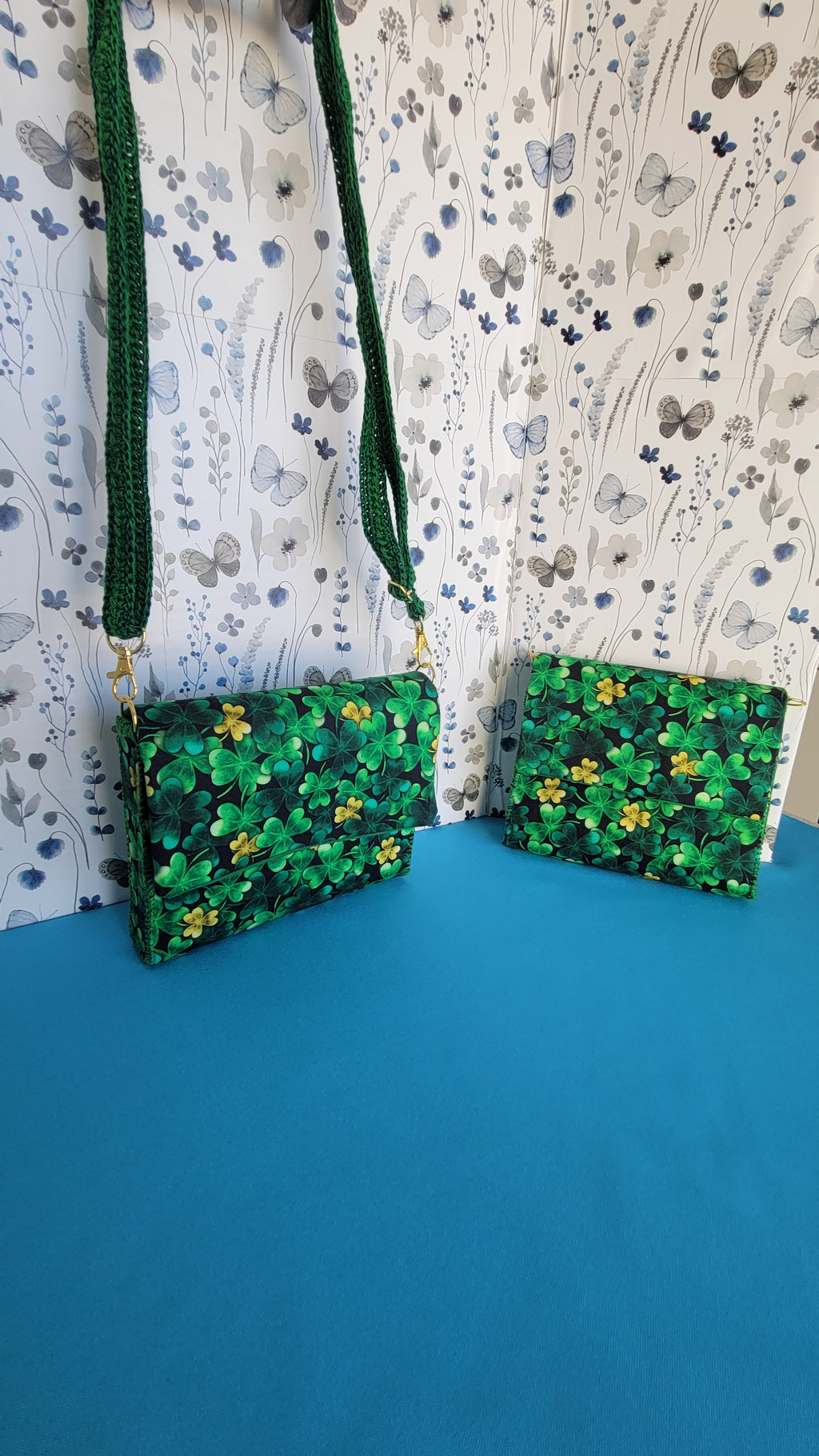 A green leaf clover fabric clutch purse hanging from it's green lace strap against a floral patterns on a blue background
