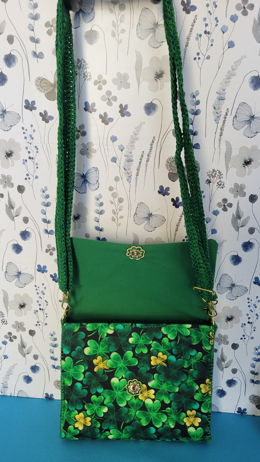 A green leaf clover fabric clutch purse hanging from it's green lace strap against a floral patterns on a blue background