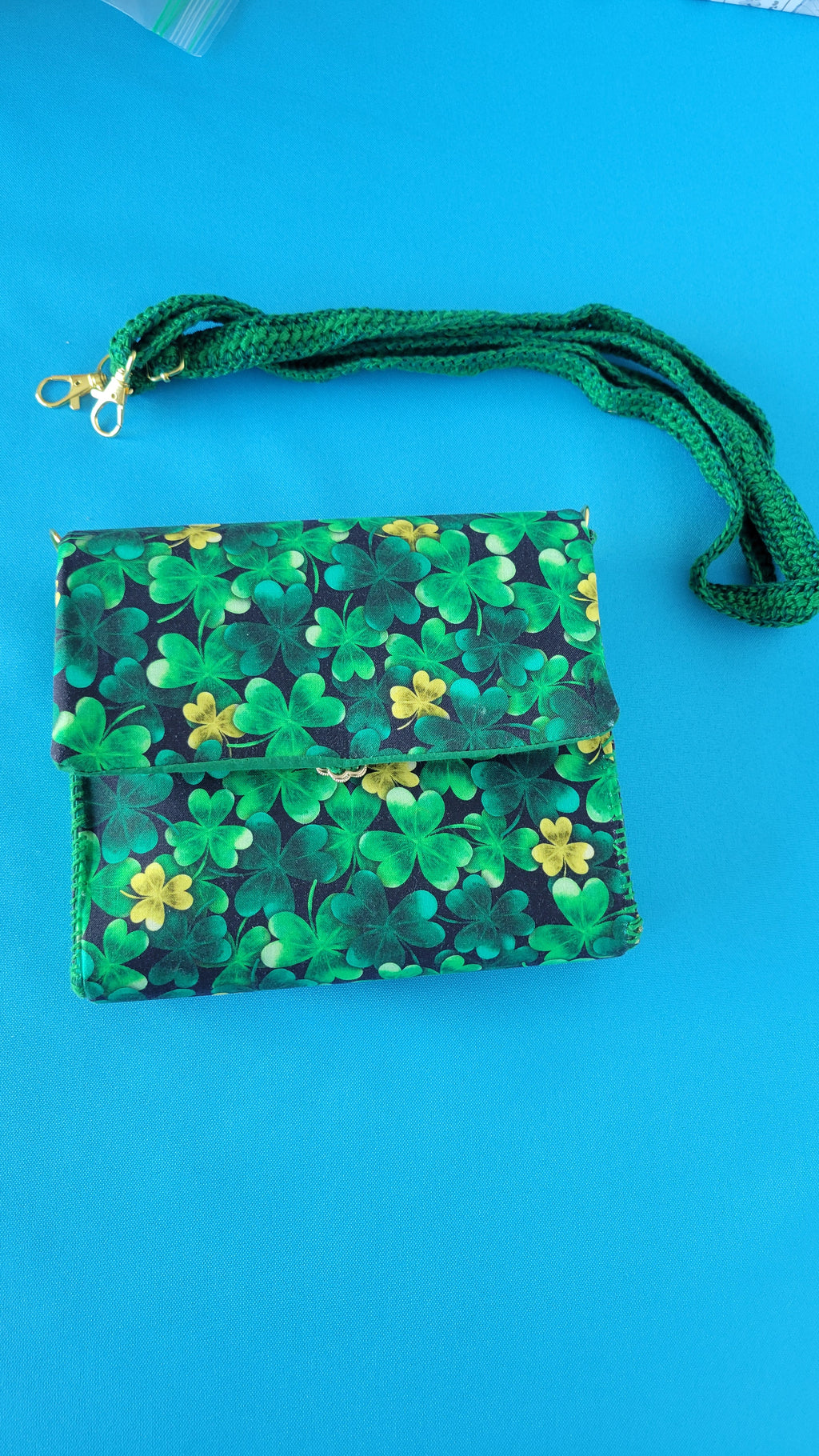 A green clutch purse with clover pattern and it handmade crochet strap on a blue background. In this picture you can clearly see the gold hooks used to attach the purse to the strap.