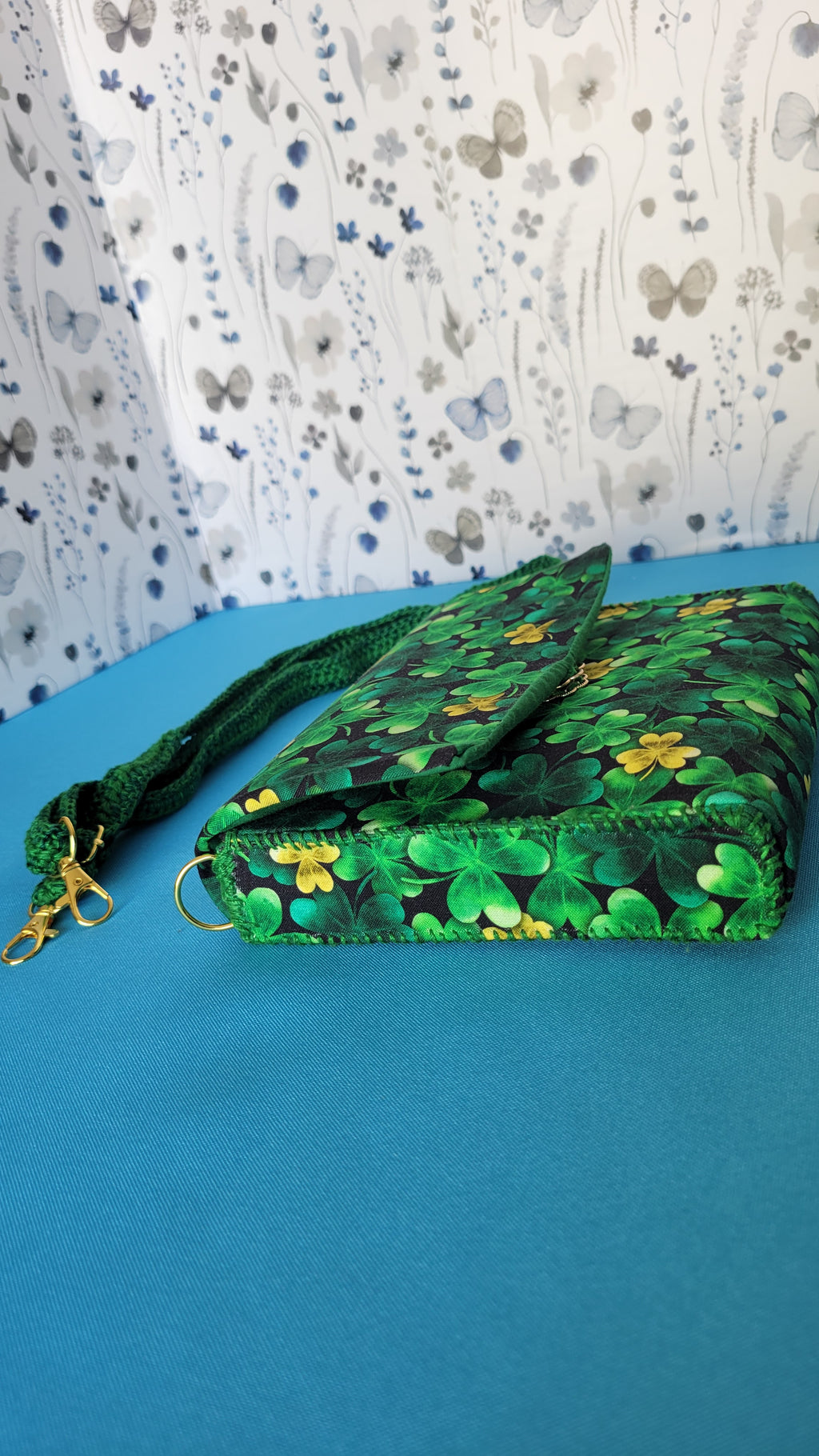 A green leaf clover fabric clutch purse. Next to the purse is it's green strap which hand crochet. They are laying on a blue background surrounded by a floral background.