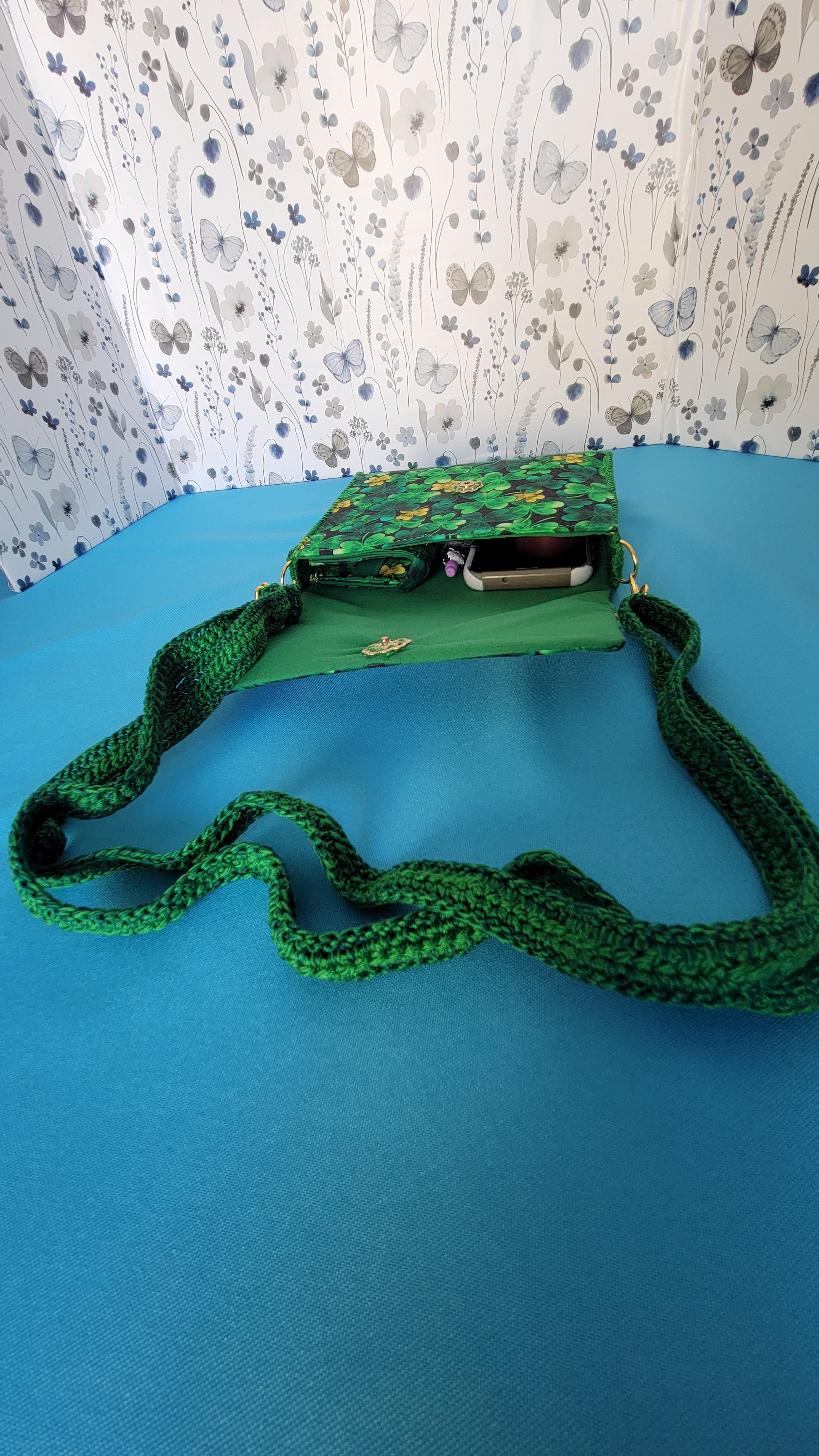 A green leaf clover fabric clutch purse with it's green lace strap laying against a blue background. You can peak inside and see ervals items it can hold. A phone, pen, wallet key and hand sanitizer. 
