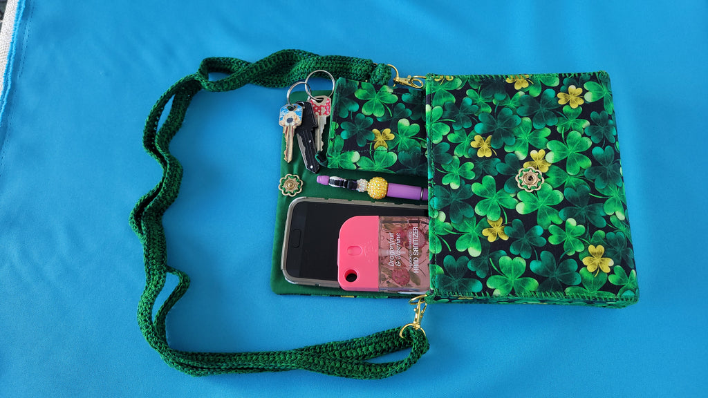 A green leaf clover fabric clutch purse with it's green lace strap laying against a blue background. The items it can fit is showed as an example of servals items this Florse purse can hold. A phone, pen, wallet, key and hand sanitizer. The closure is a vintage flower which can be clearly seen in this photo.