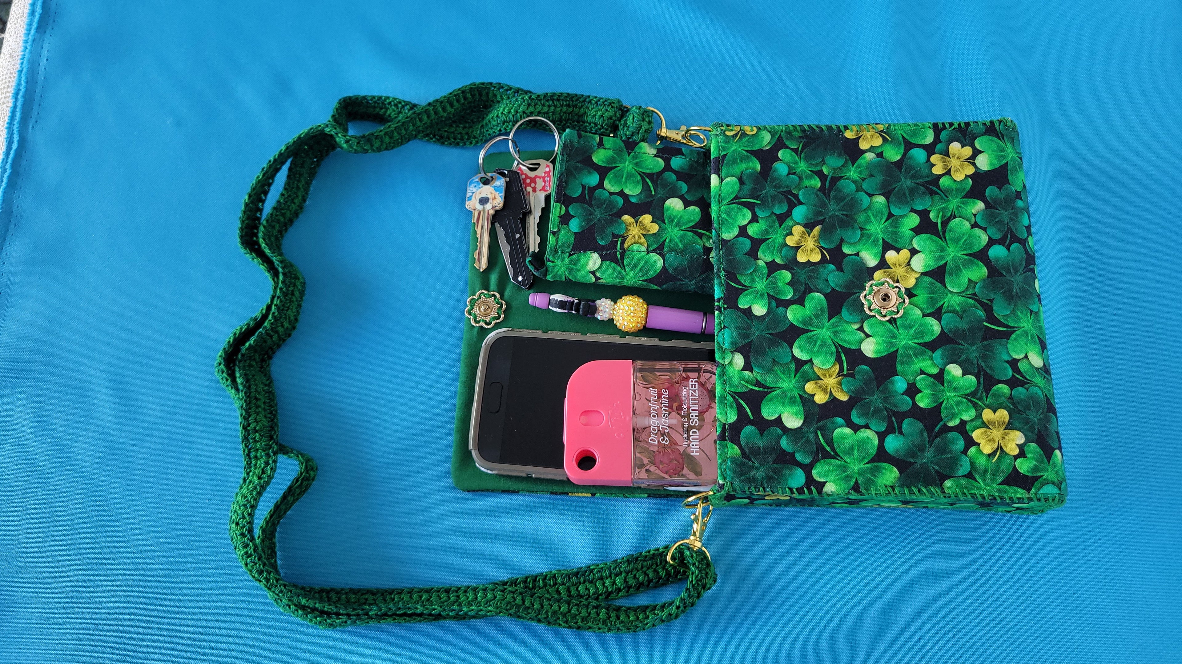 A green leaf clover fabric clutch purse with it's green lace strap laying against a blue background. The items it can fit is showed as an example of servals items this Florse purse can hold. A phone, pen, wallet, key and hand sanitizer. The closure is a vintage flower which can be clearly seen in this photo.