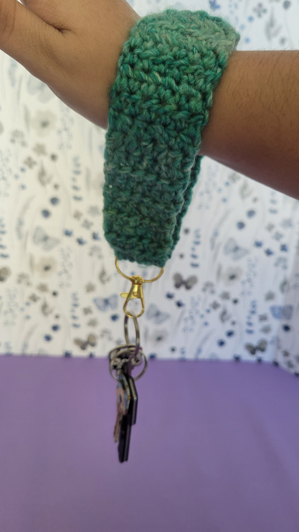 Green crochet key bracelet item with a key attached, held by a person against a textured background. The size of the bracelet is 5 inches.